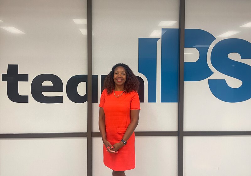 A woman wearing a bright orange dress stands in front of a wall with the words "Team IPS."