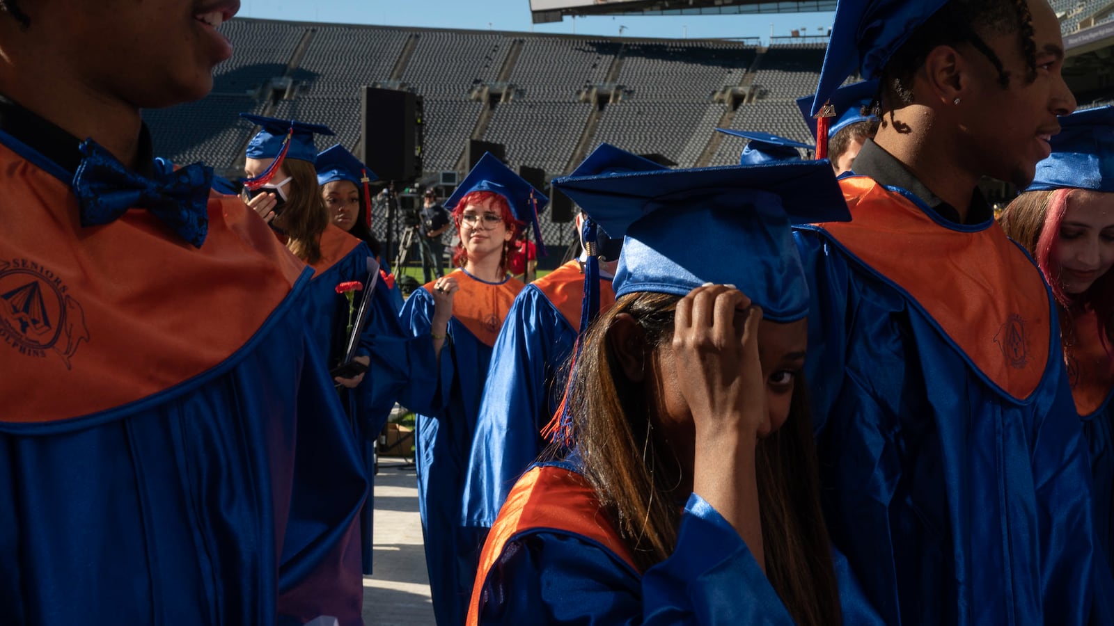 Several recently graduated students wearing blue and orange caps and gowns make their way off of a stadium field at the conclusion of their ceremony.