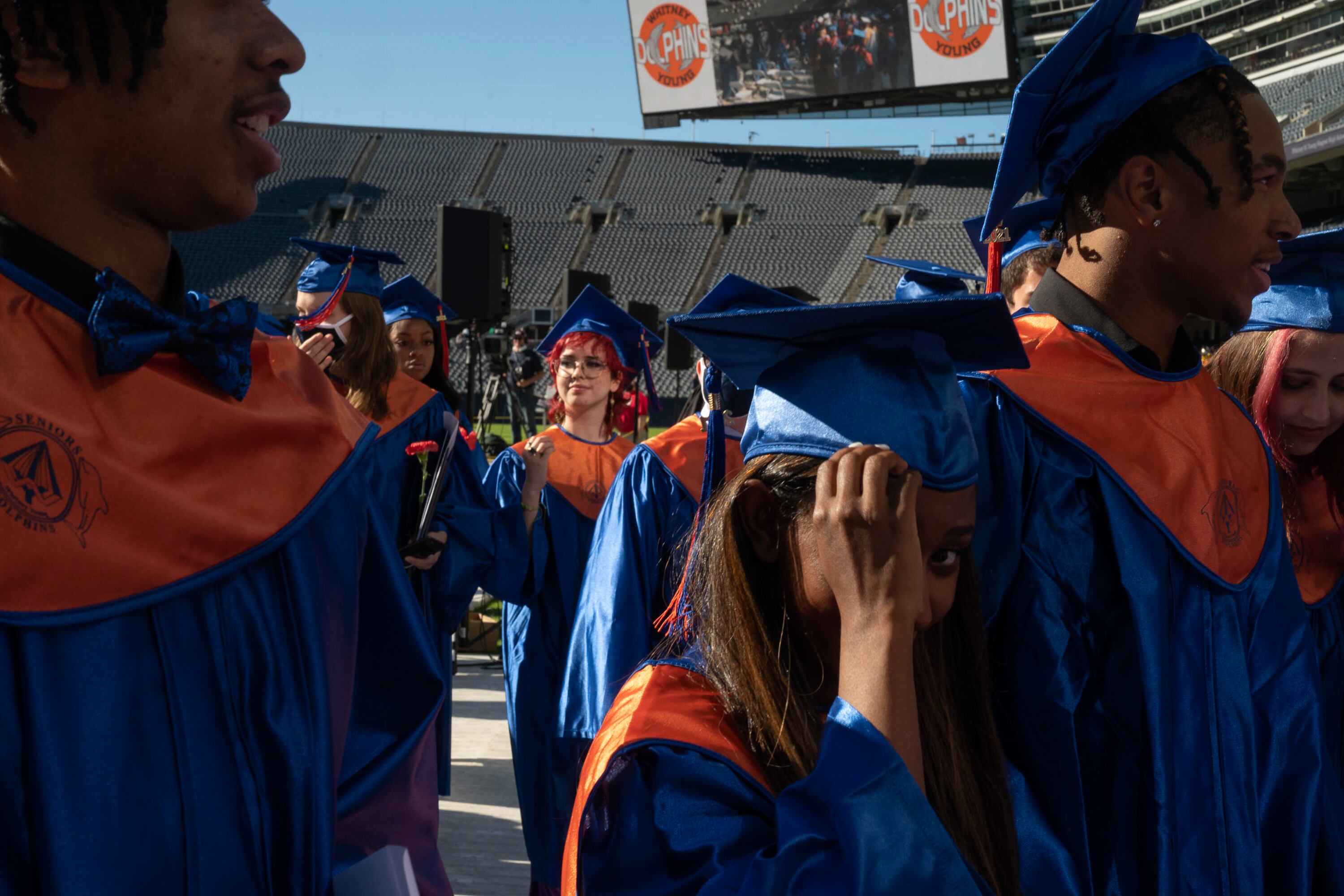 Several recently graduated students wearing blue and orange caps and gowns make their way off of a stadium field at the conclusion of their ceremony.