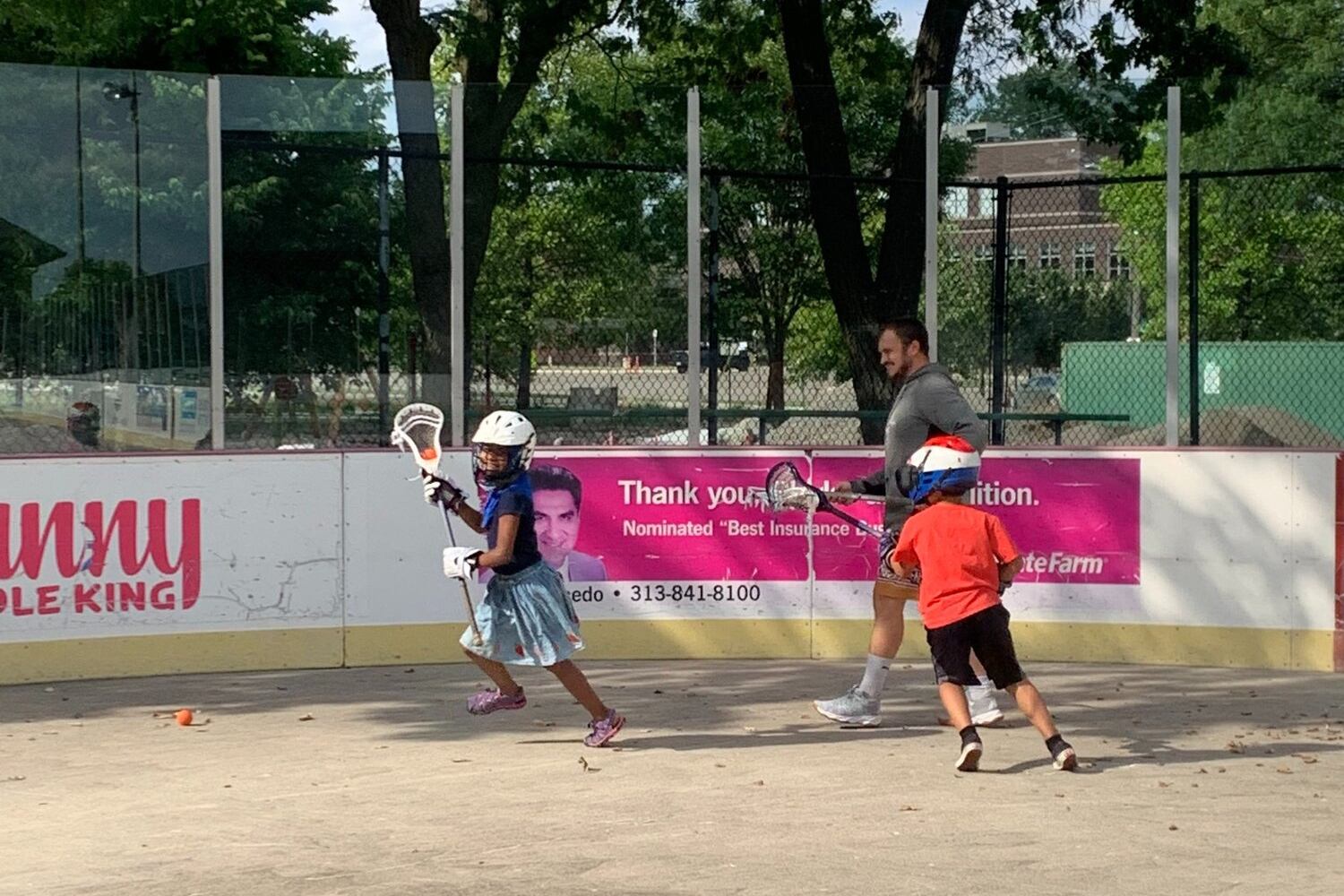 Two kids holding lacrosse sticks play, with an adult male looking on, at a park in Detroit.