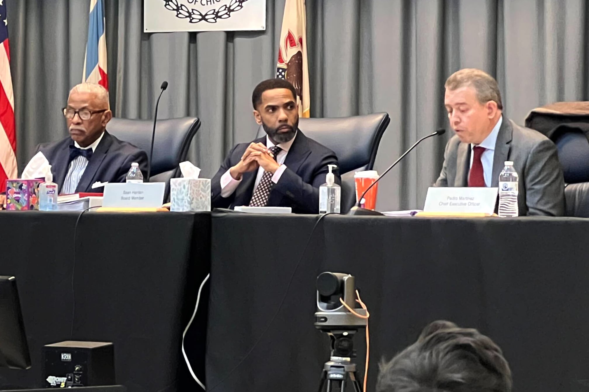 Three men in dark suits sit at a long podium.