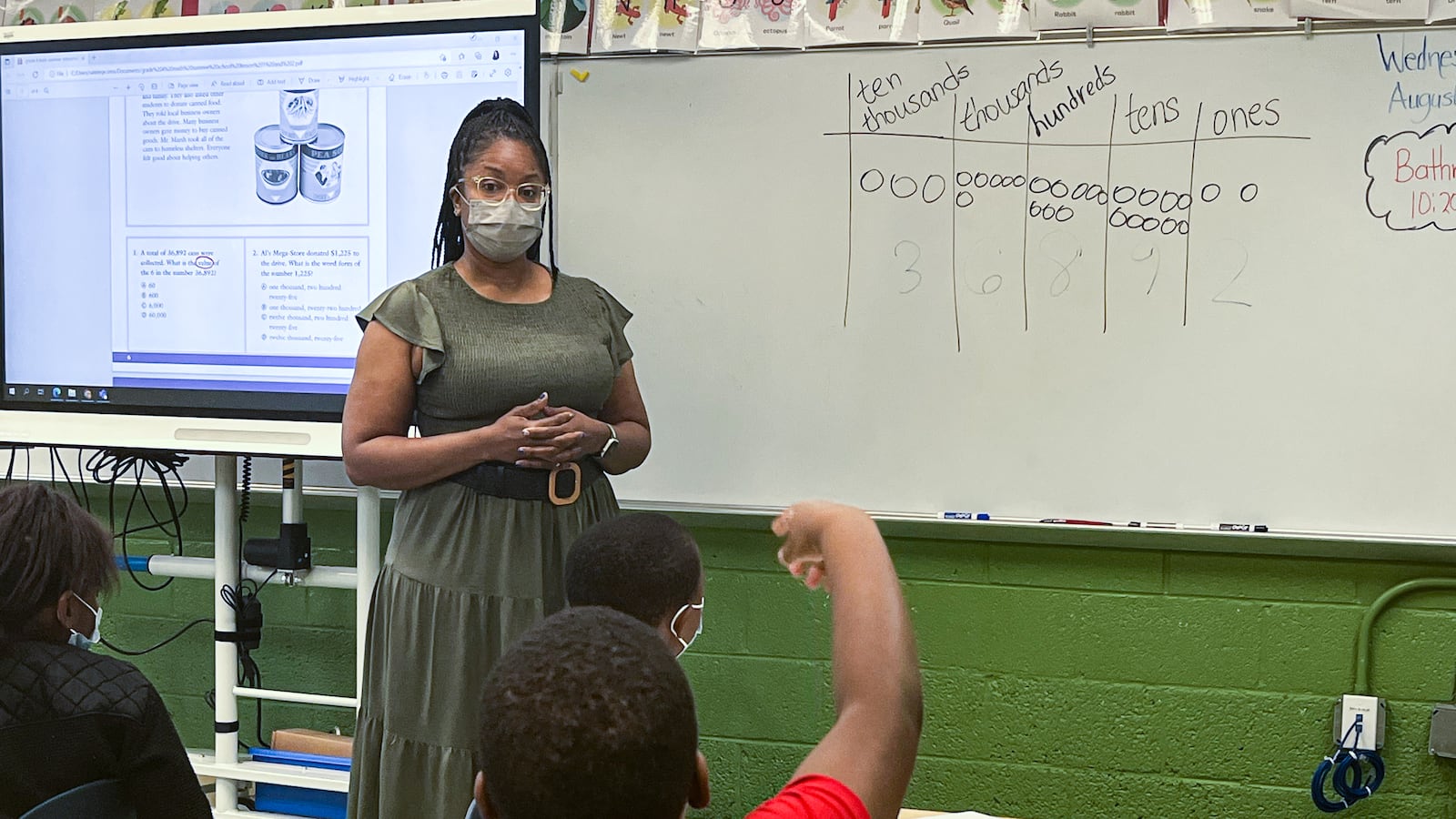 A teacher wearing a mask stands at the front of a classroom in front of a whiteboard that shows mathematical principles. In the foreground, a student in a red shirt has his hand raised.