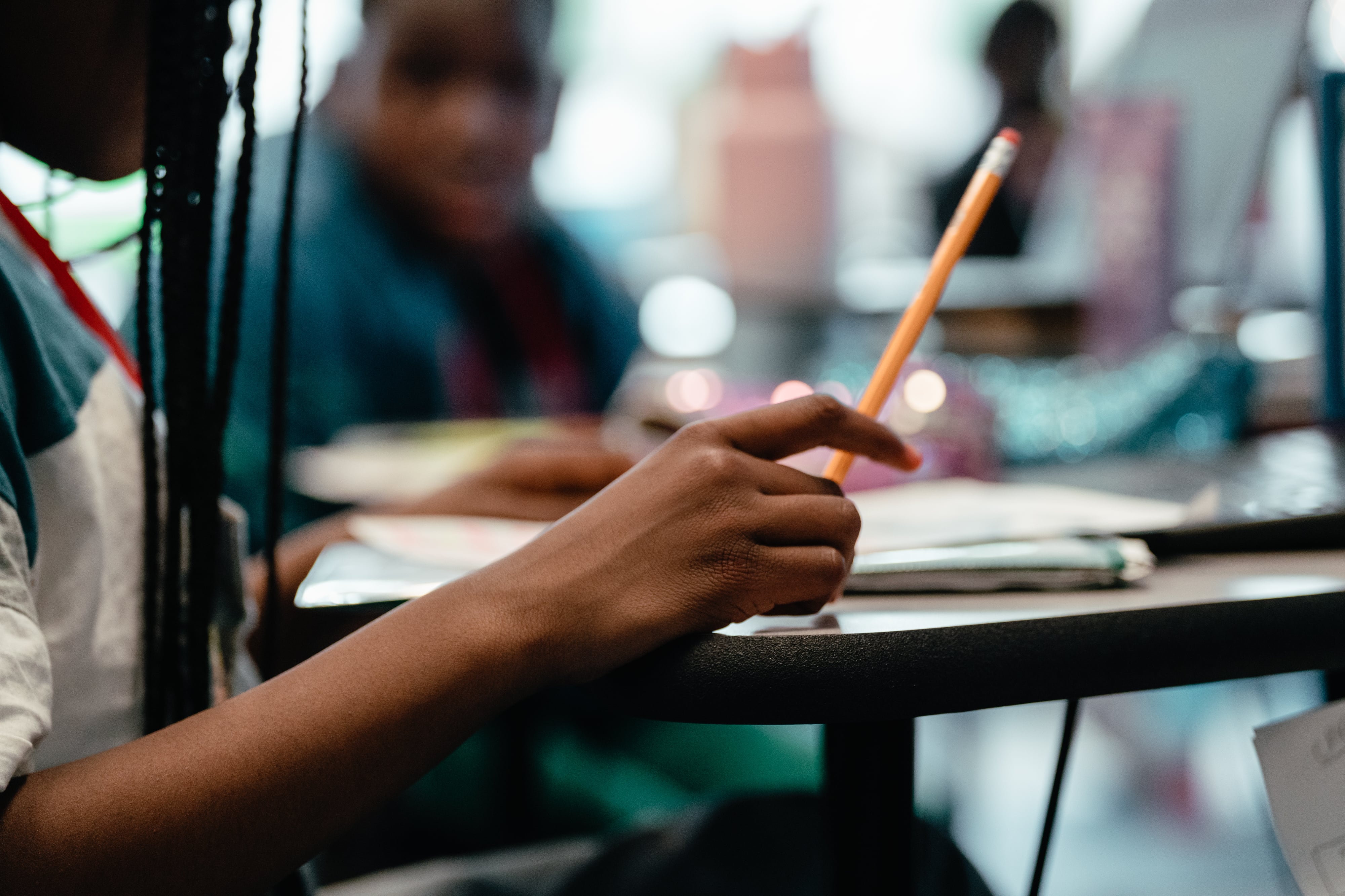 A student holds a yellow #2 pencil in the air above a desk.