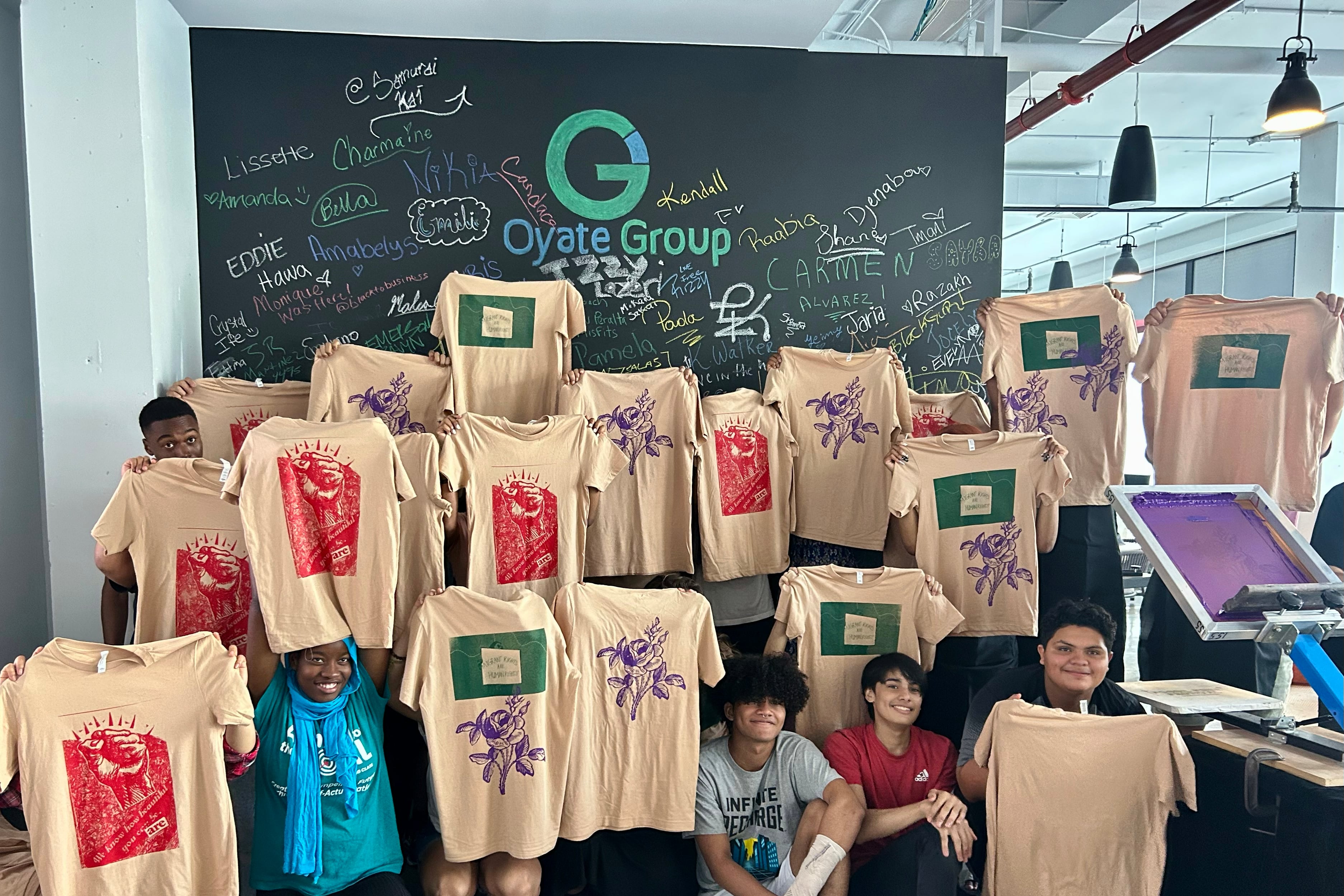 People holding up beige T-shirts in a room with a green chalkboard.