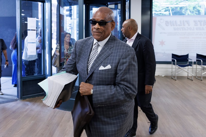 A Black man in sunglasses and a suit walks in a building holding a stack of papers with a person in the background.