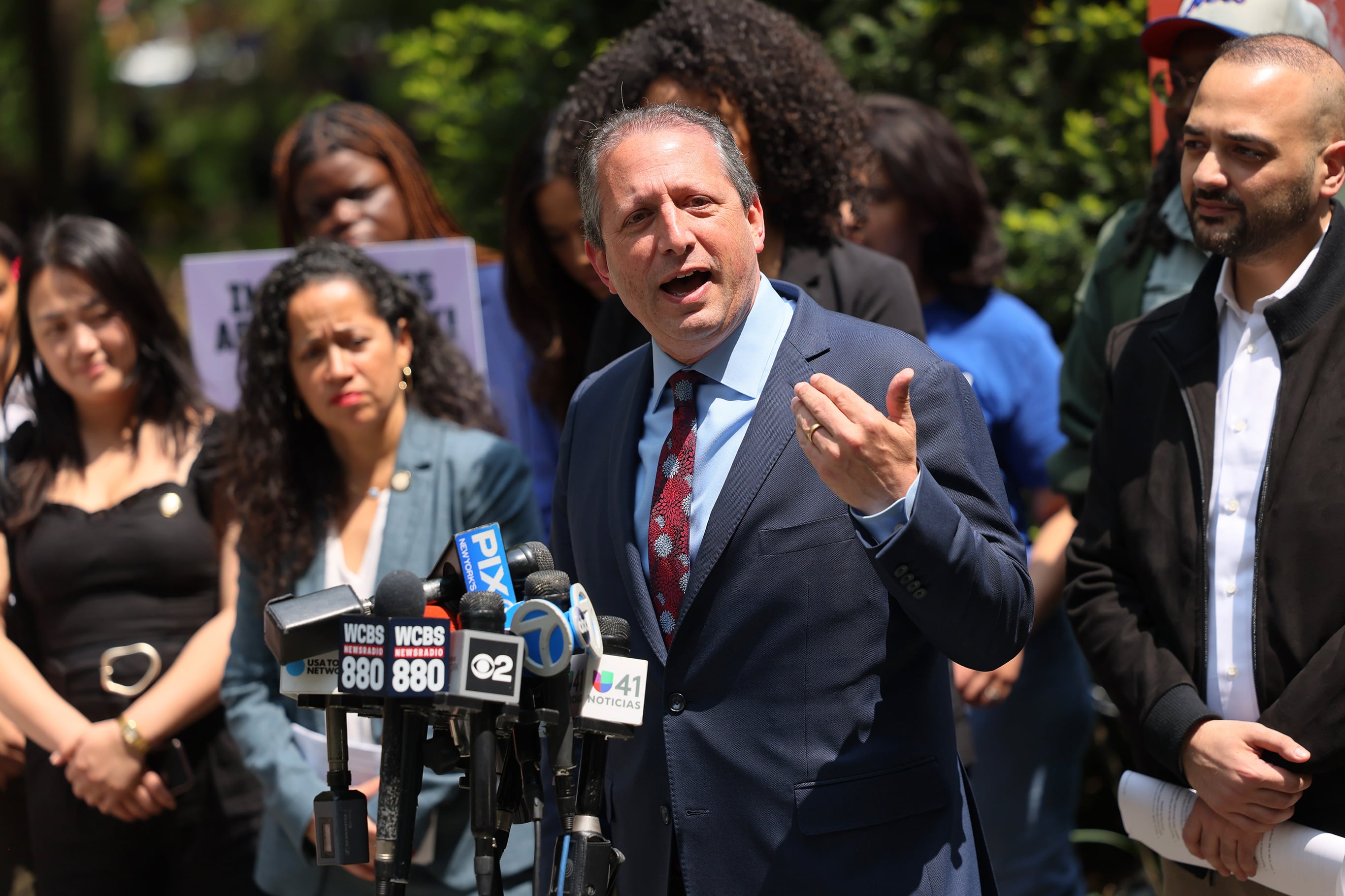 A man wearing a dark suit with a red tie speaks at a podium outside with several people standing behind him.