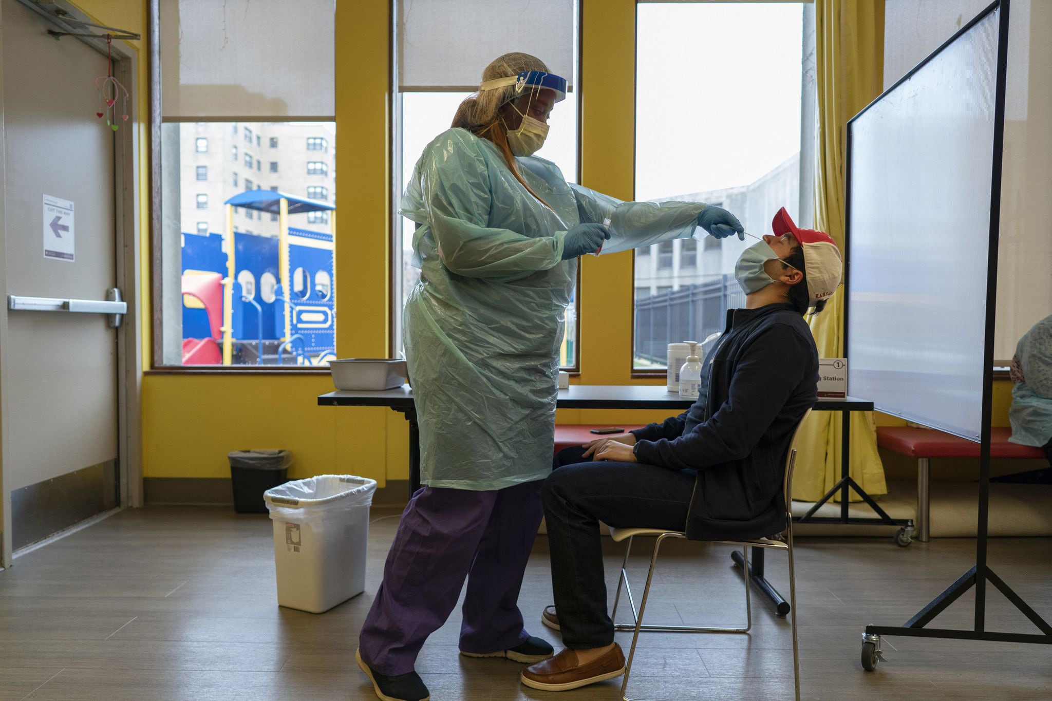 A person wearing scrubs and a face shield tests a man sitting in a chair for COVID-19.