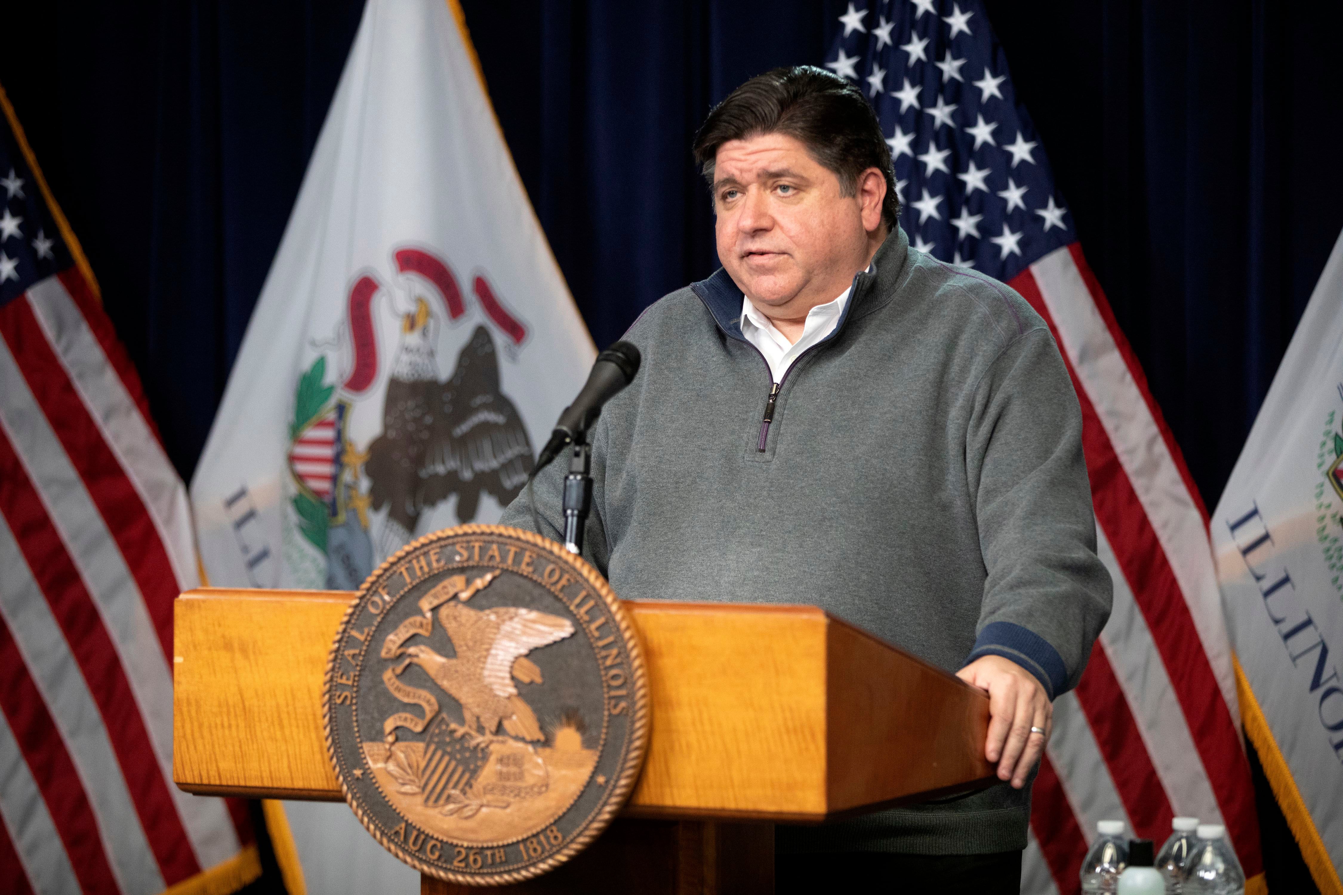 Illinois Governor J.B. Pritzker stands at a podium, wearing a grey pull-over sweater.
