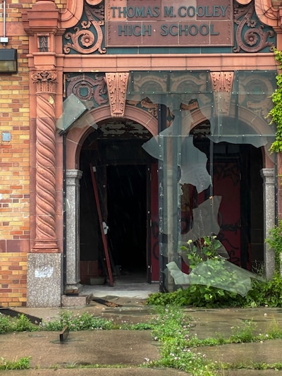 A photograph of the damaged entrance to Cooley High School, a brown brick building with shattered glass panels.