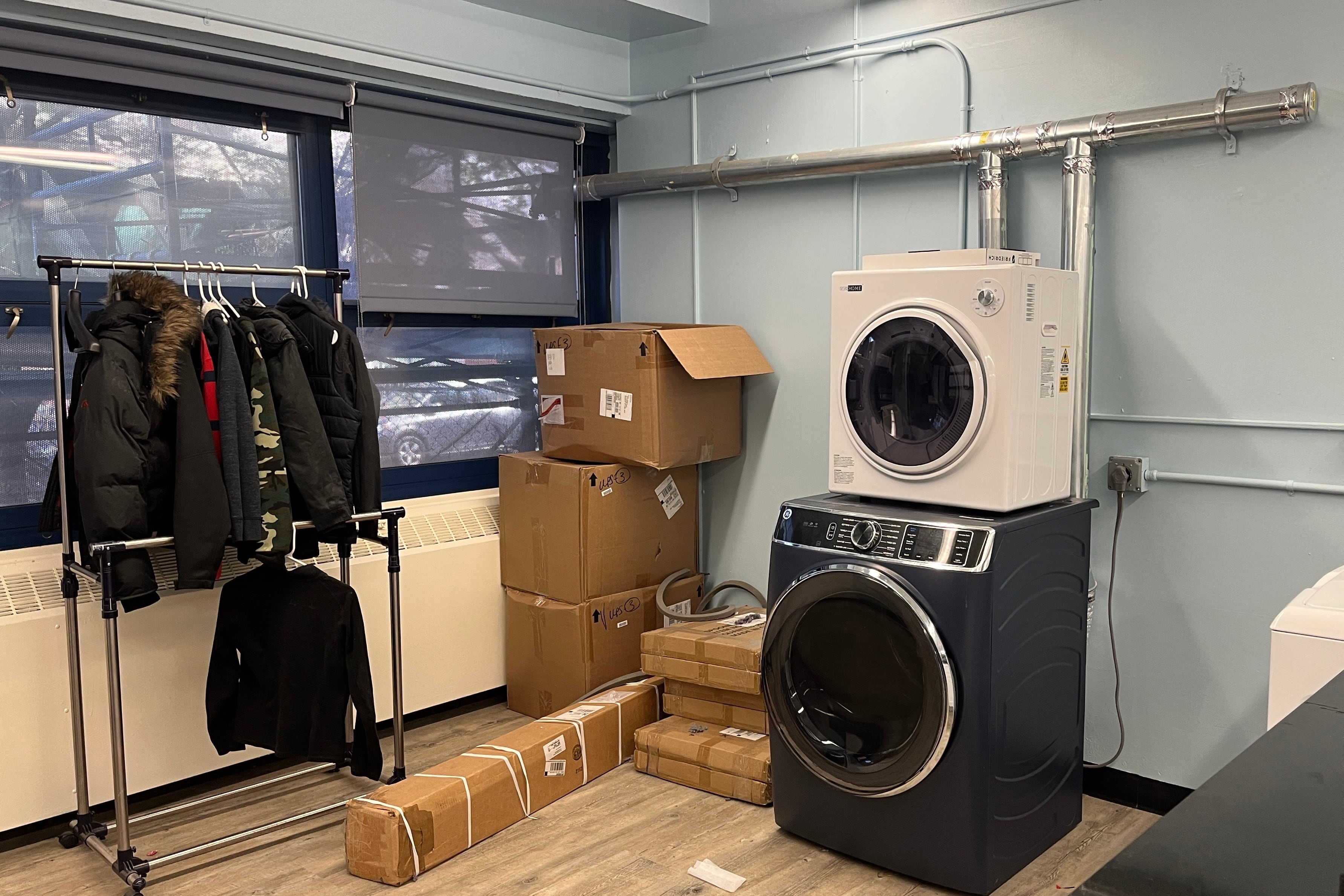 A room with windows on one wall, a rack of clothing some cardboard boxes and a washer and dryer.