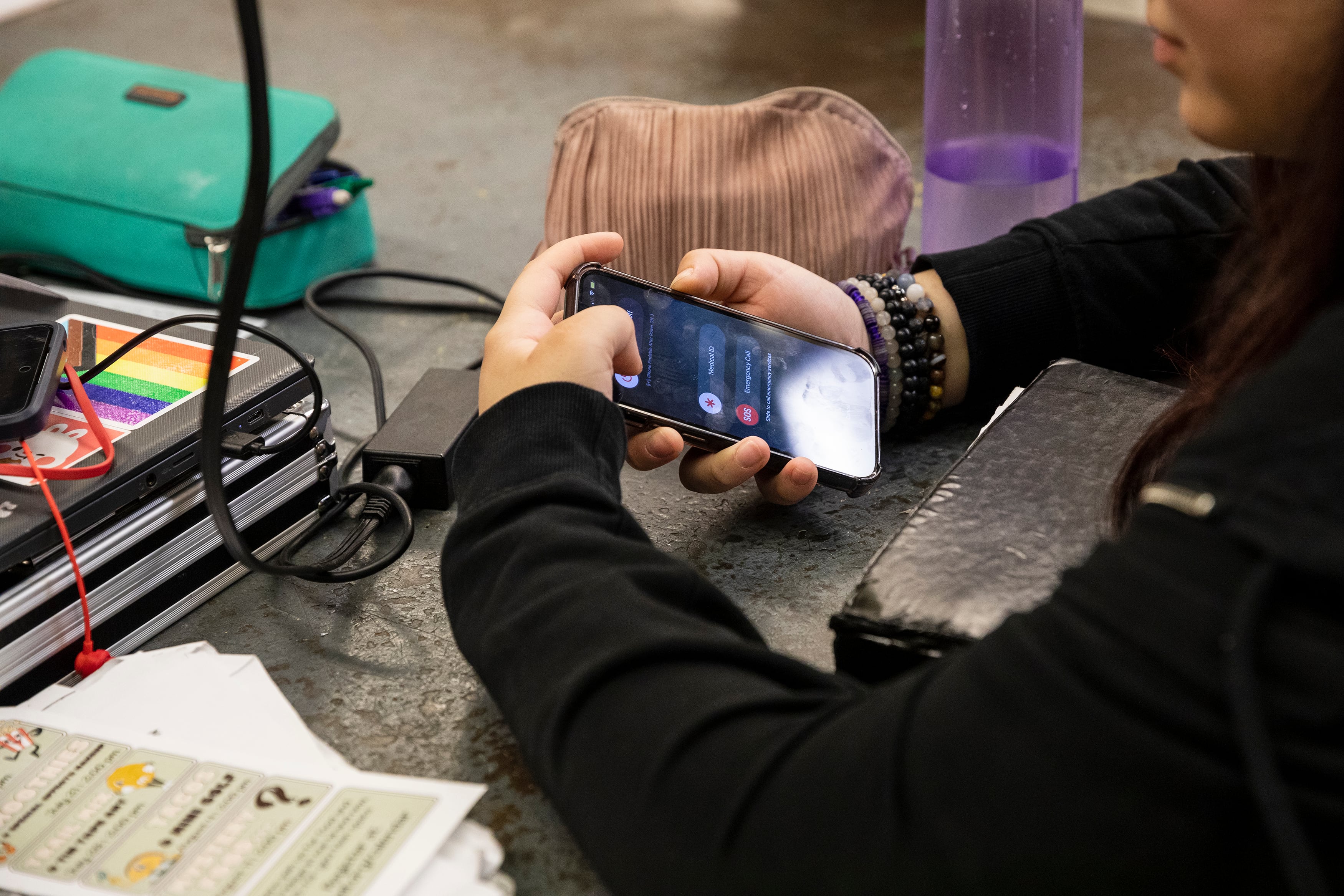 A middle school student holds her cellphone at a desk surrounded by her books, pencils and a water bottle in class.