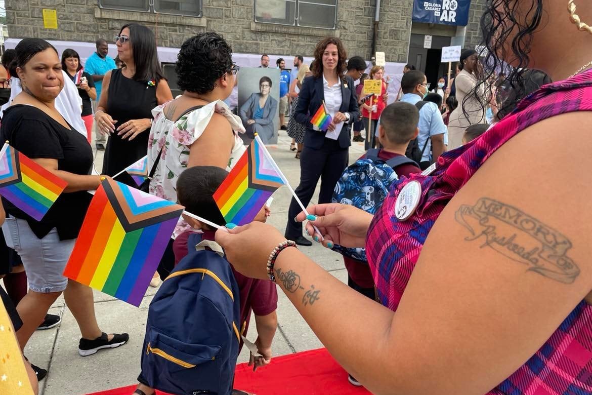 A small crowd of students and parents hold small pride flags inside an elementary school.