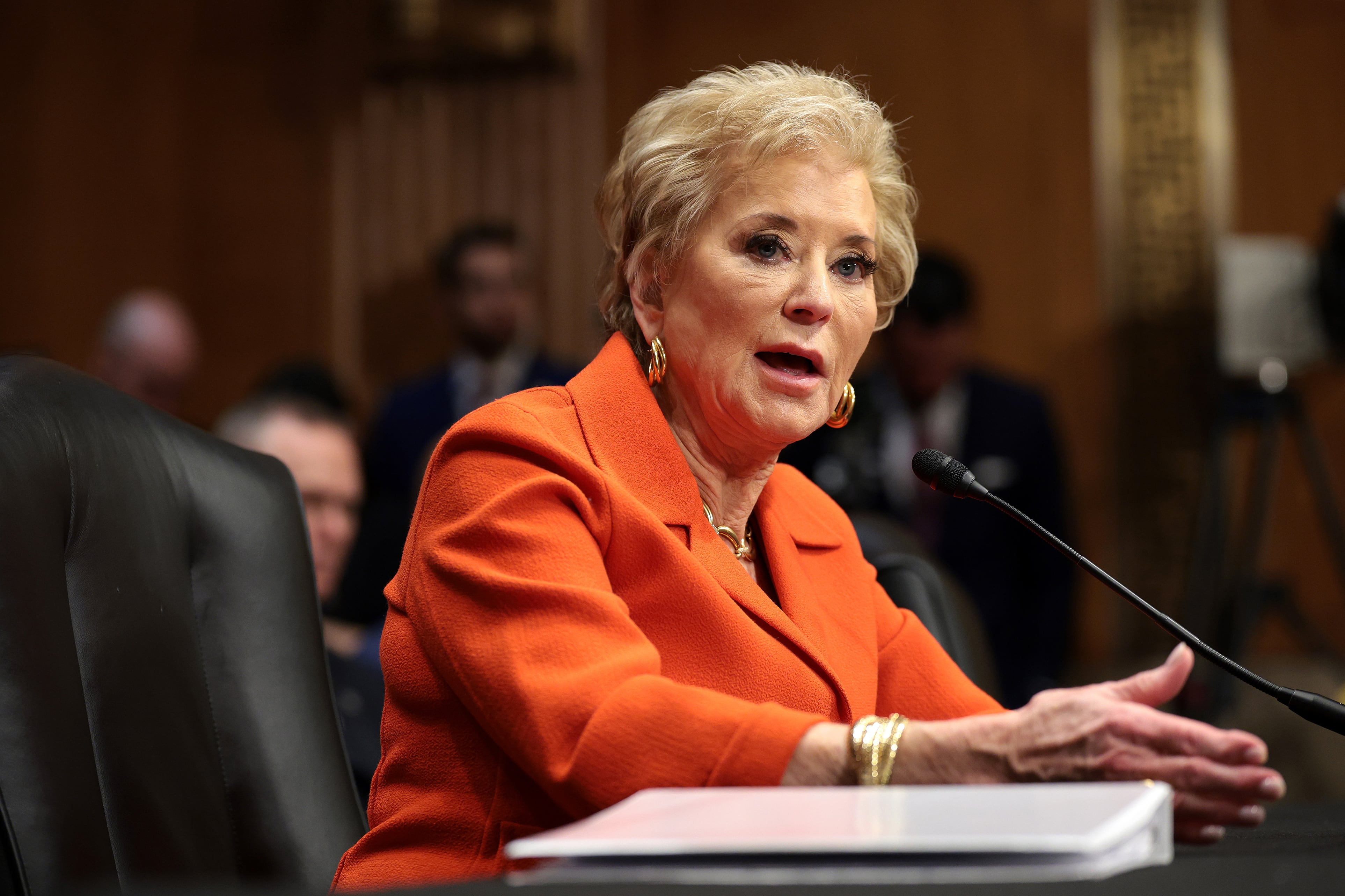 A woman with short blonde hair and wearing a bright orange suit sits in front of a microphone.