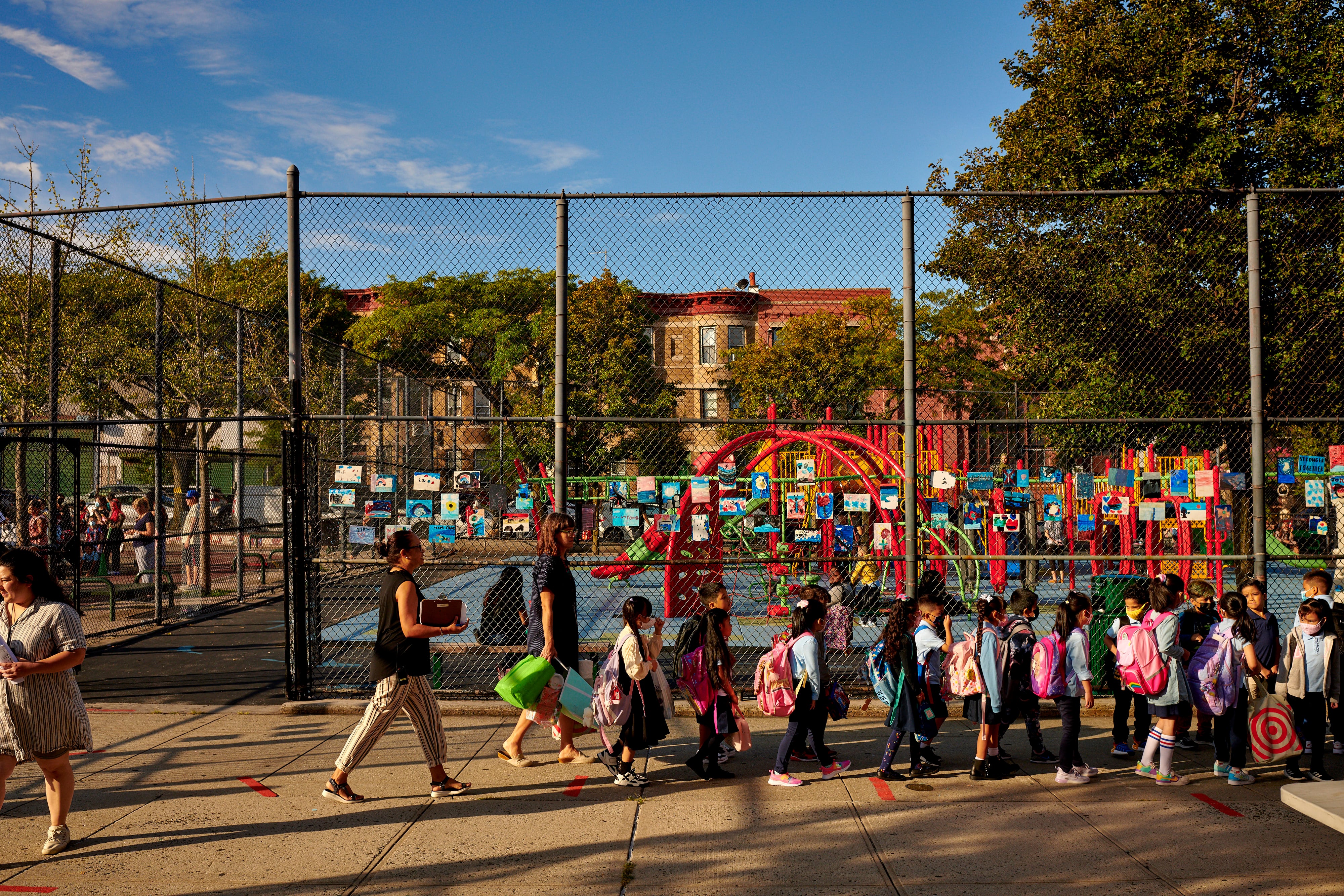 Children with backpacks walk in front of a school playground followed by their teacher.