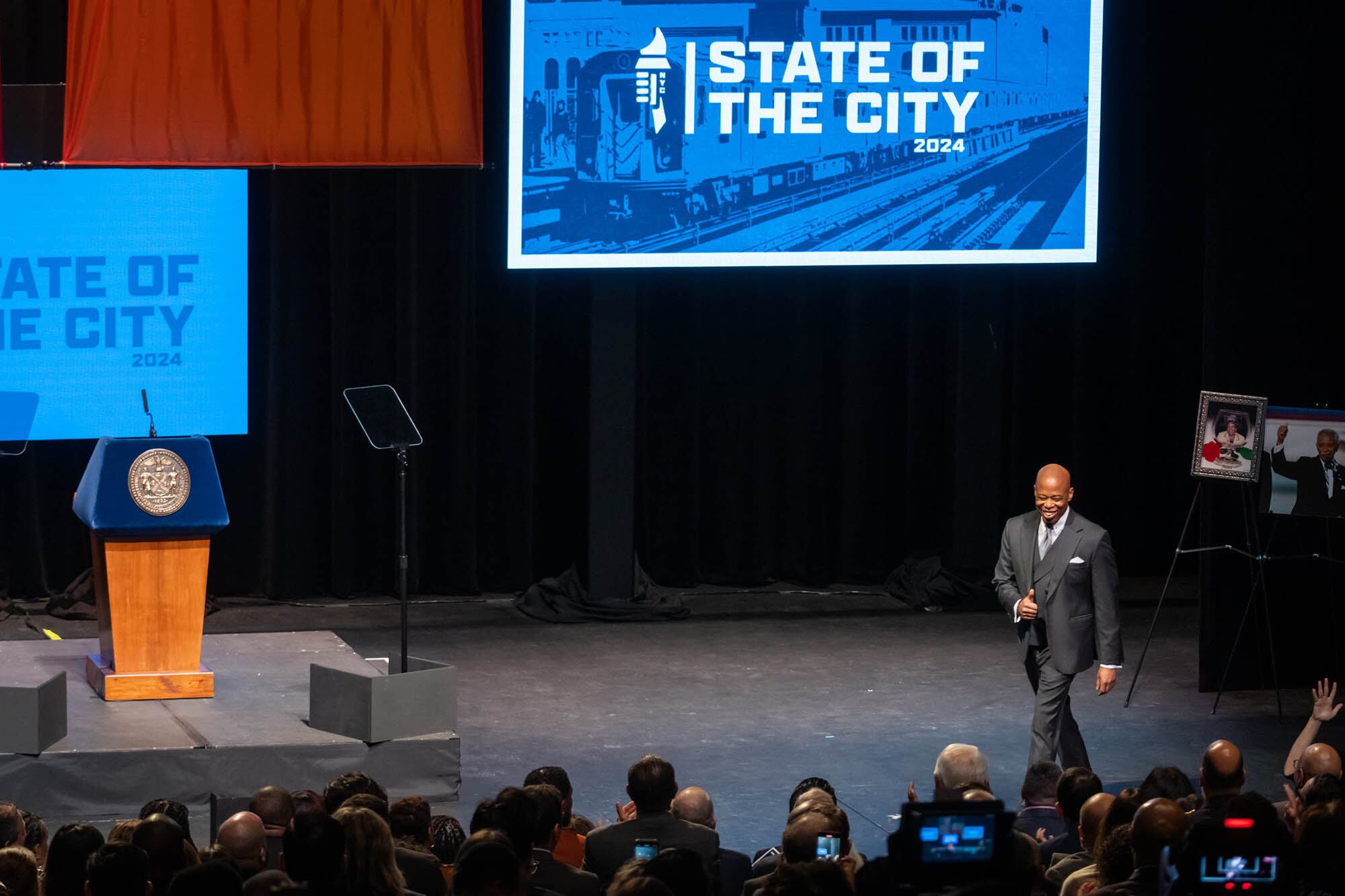 A man wearing a dark suit walks across a stage with people sitting in chairs in the foreground and a large blue screen that reads "State of the City."