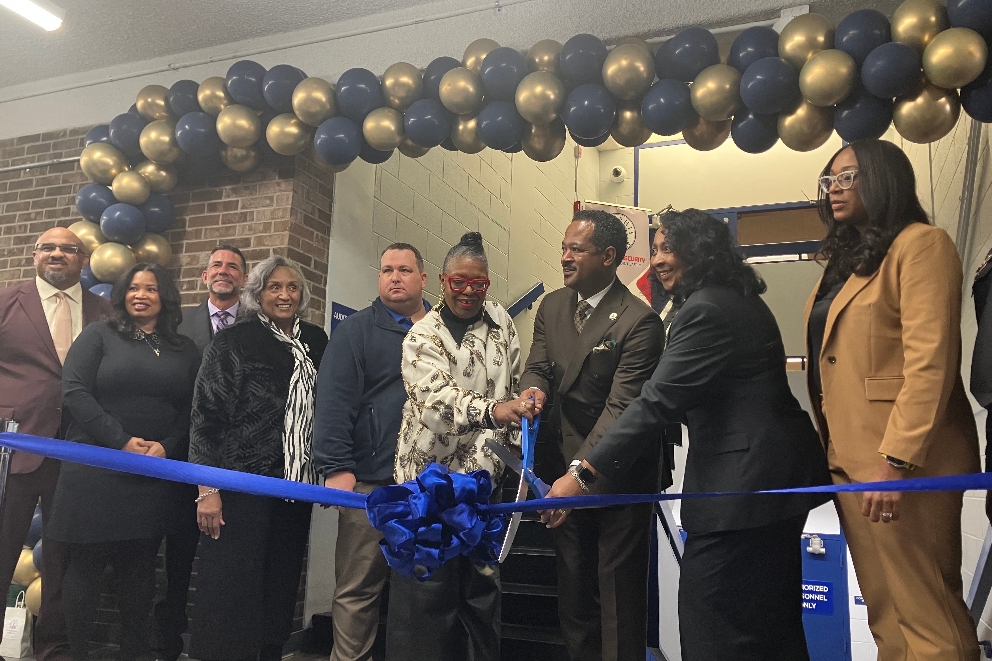 Nine people stand behind a bright blue ribbon with a big bow, under a blue and gold balloon arch. Two people in the center, a man and a woman, hold a big pair of blue scissors.