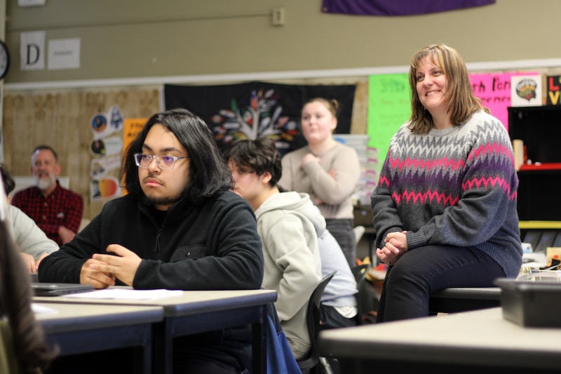 A teacher with short blonde hair sits on a desk while a couple other students sit at their desks in a classroom.
