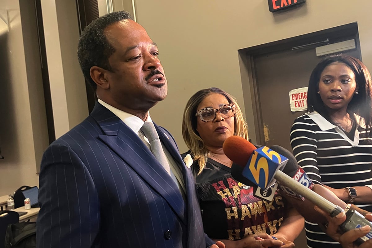 A man in a dark suit jacket speaks to a cluster of microphones.