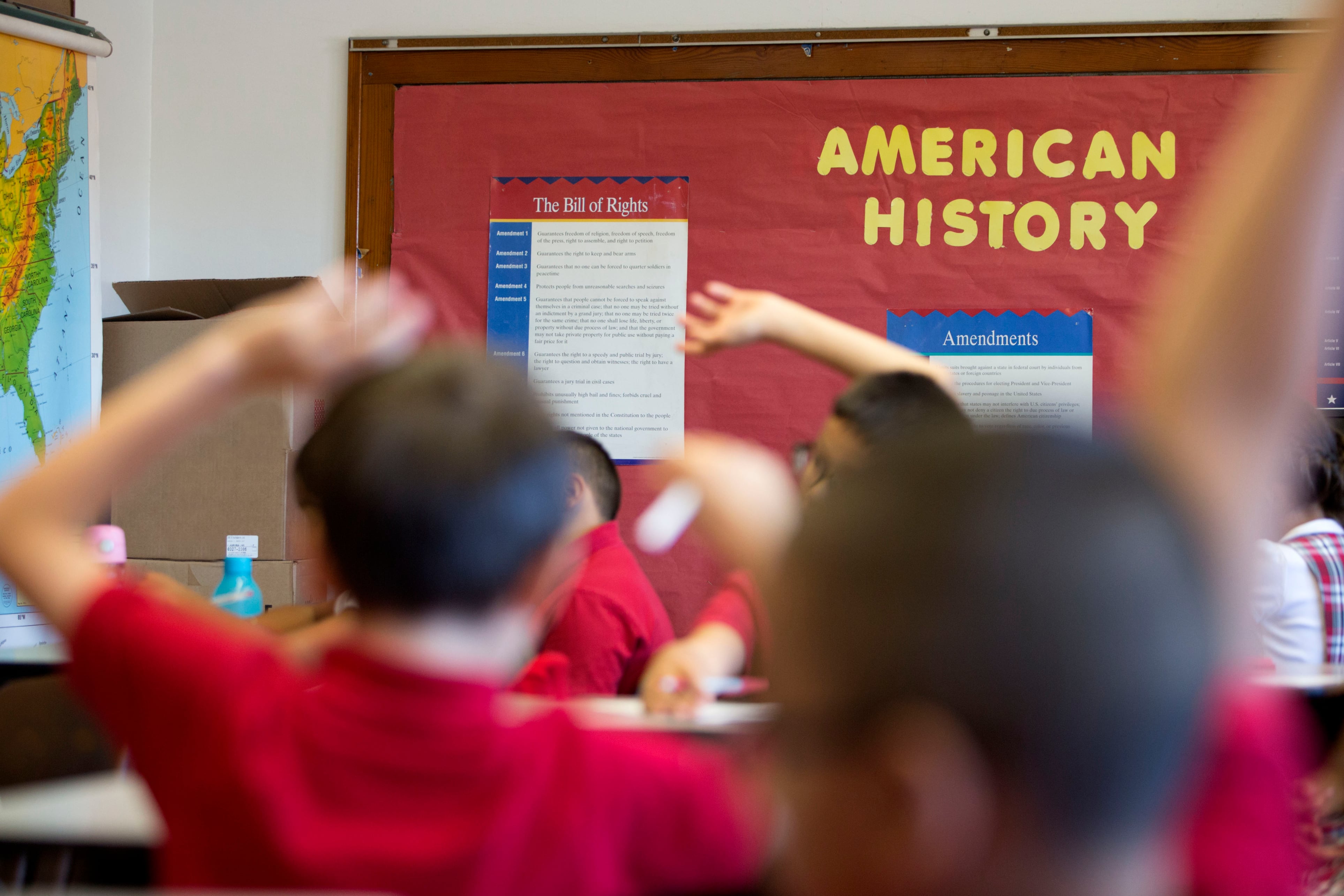 Students raise their hands in a classroom with a bulletin board that says “American History.”