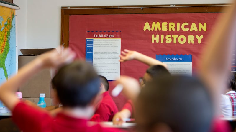 Students raise their hands in a classroom with a bulletin board that says “American History.”