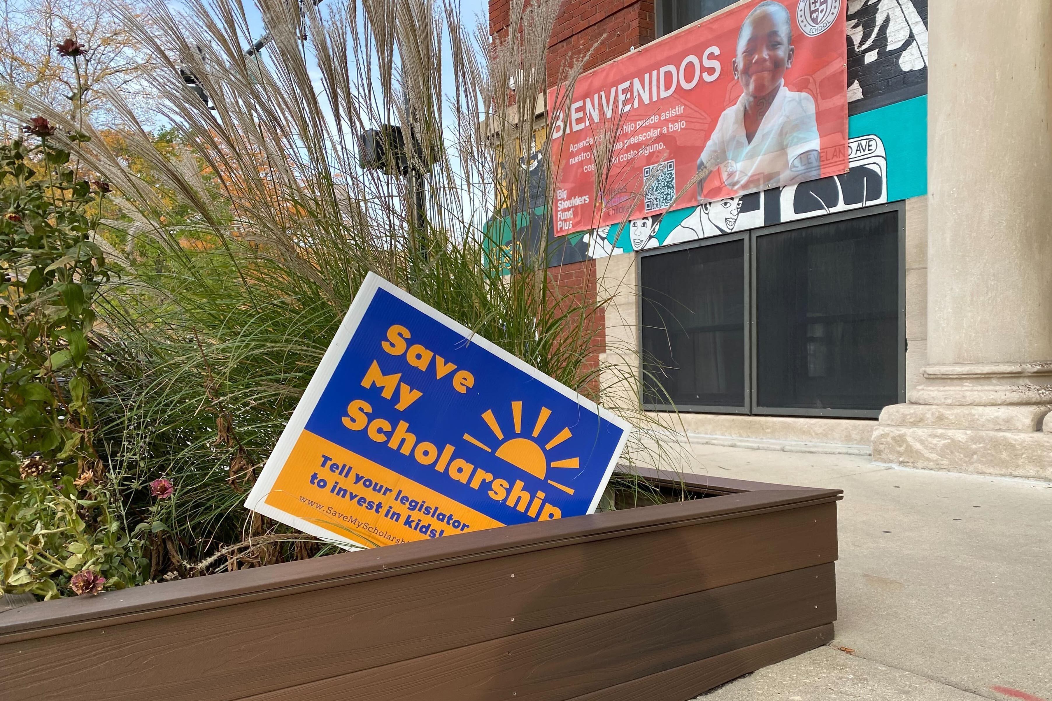 A blue and yellow sign that reads" save my scholarship" sits in a flower bed in front of a school building.