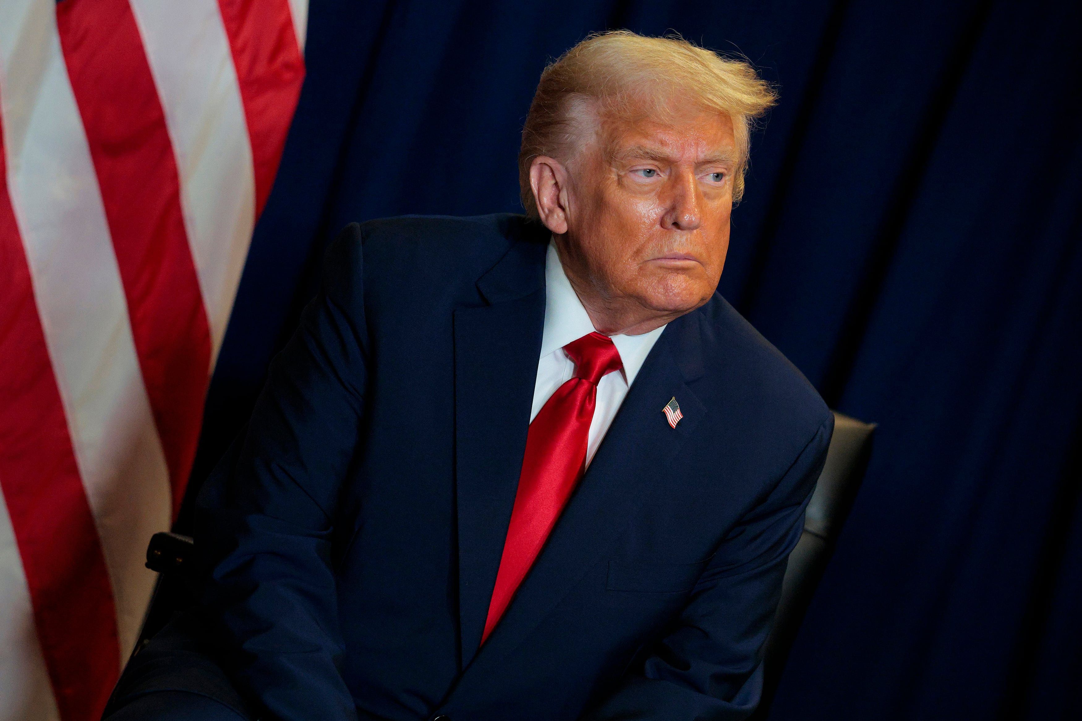 A photograph of President Donald Trump, a white man with yellow hair and wearing a dark suit and a red tie sitting in front of a dark background with part of the American flag on his left side.