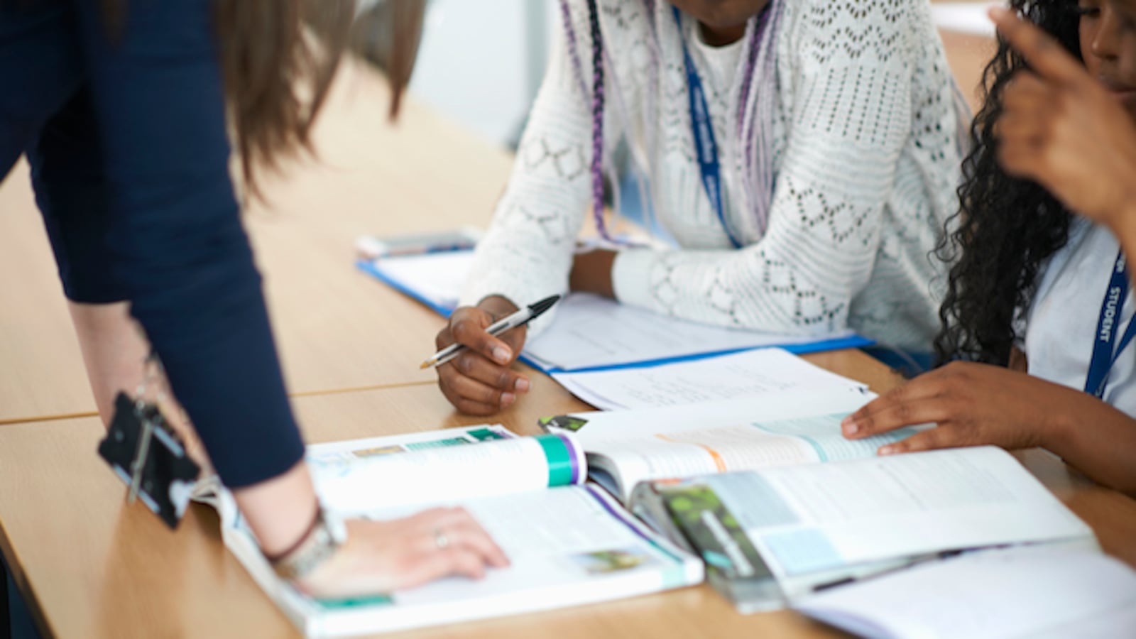 Close-up of the arms of a teacher and student facing each other opposite a desk with workbooks on it.