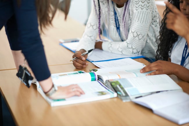 Close-up of the arms of a teacher and student facing each other opposite a desk with workbooks on it.