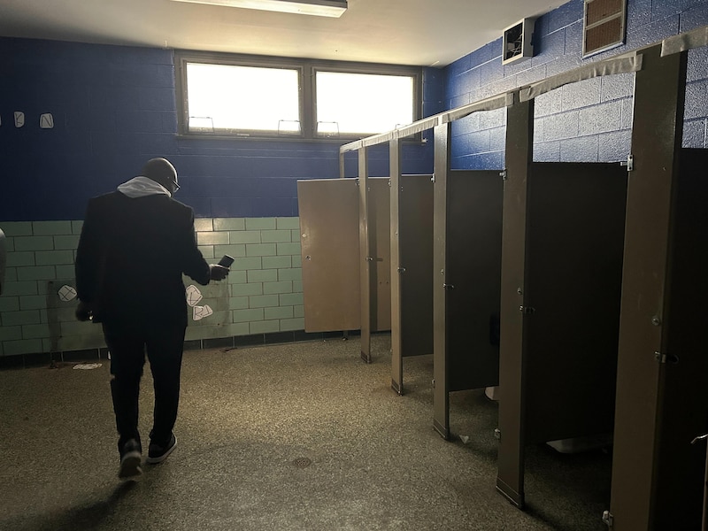 A man wearing a dark jacket holds a phone up to illuminate an empty school bathroom with stalls on the right side of the room.