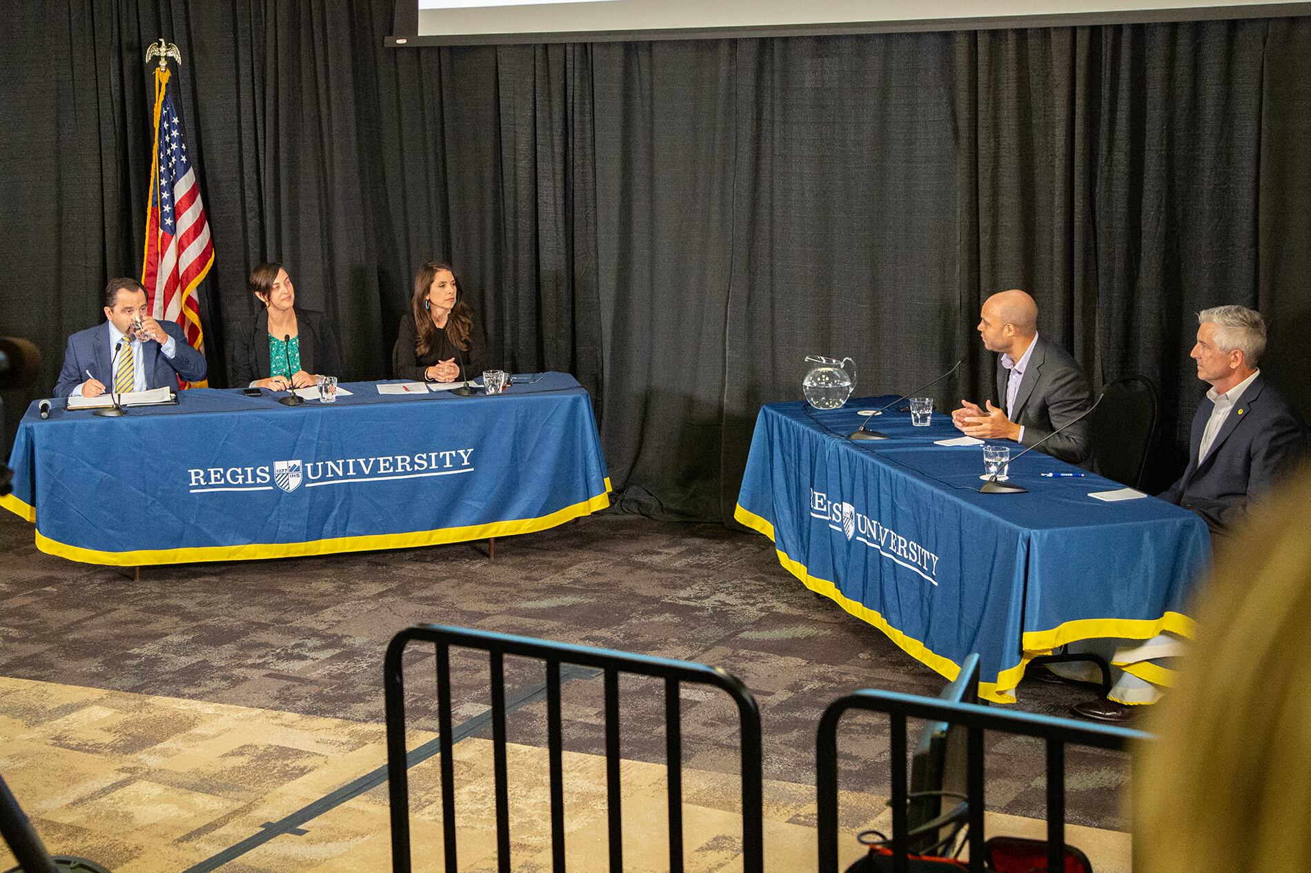 Five people sit at tables draped with banners that say Regis University. Three are moderators. Two are Denver school board candidates Kwame Spearman and John Youngquist.