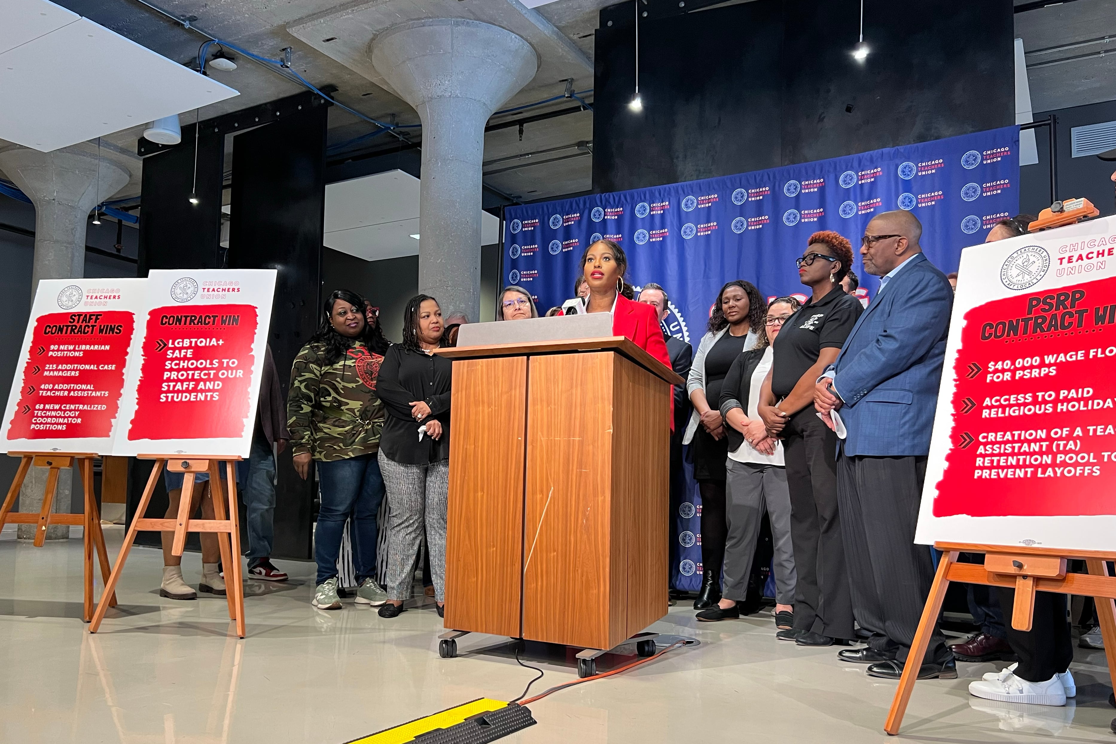 A Black woman wearing a red suit jacket speaks from behind a wooden podium and in front of a blue background an a group of people stand next to her.