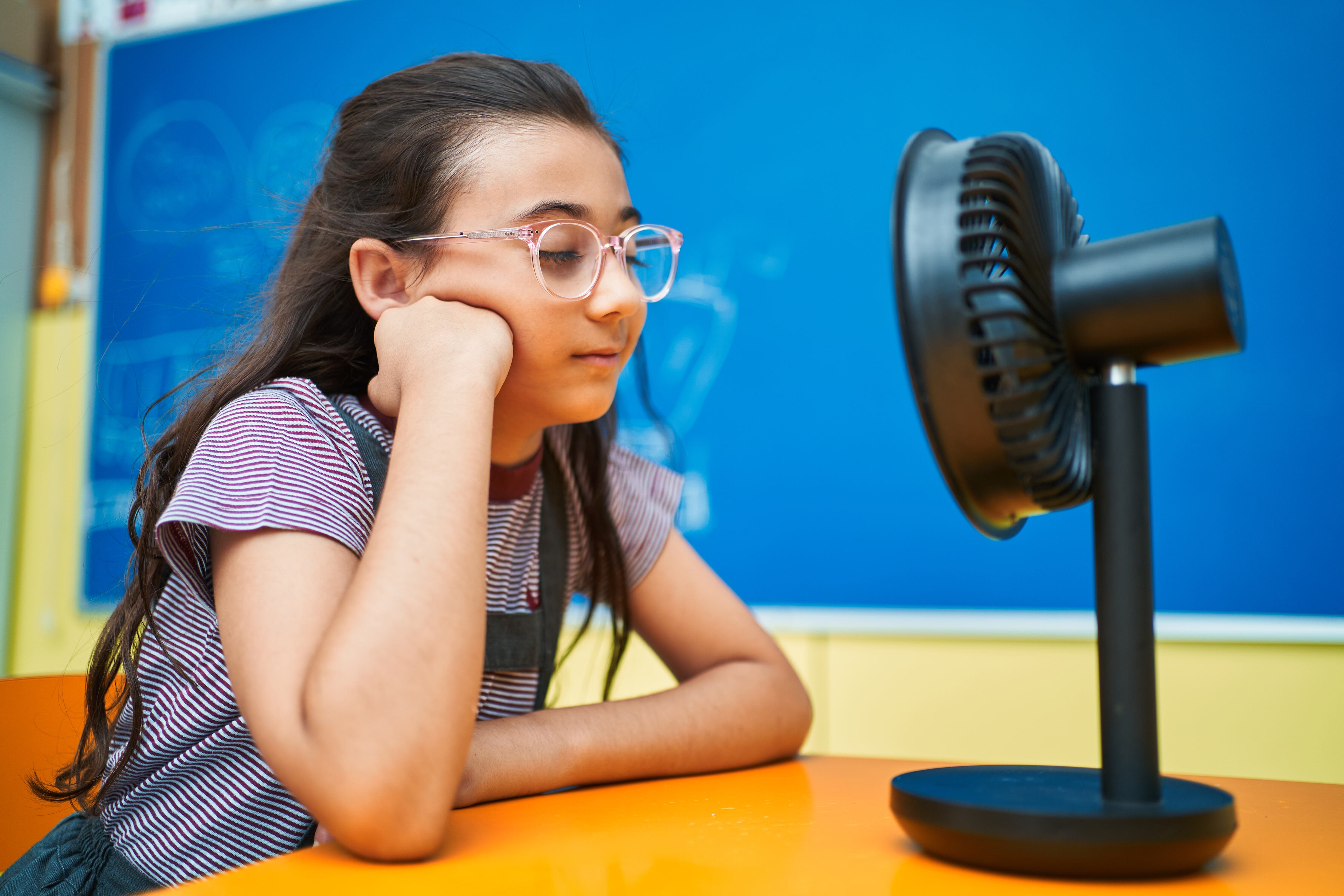Young student with long dark hair sits facing a small fan that is sitting on her desk with a blue wall in the background.
