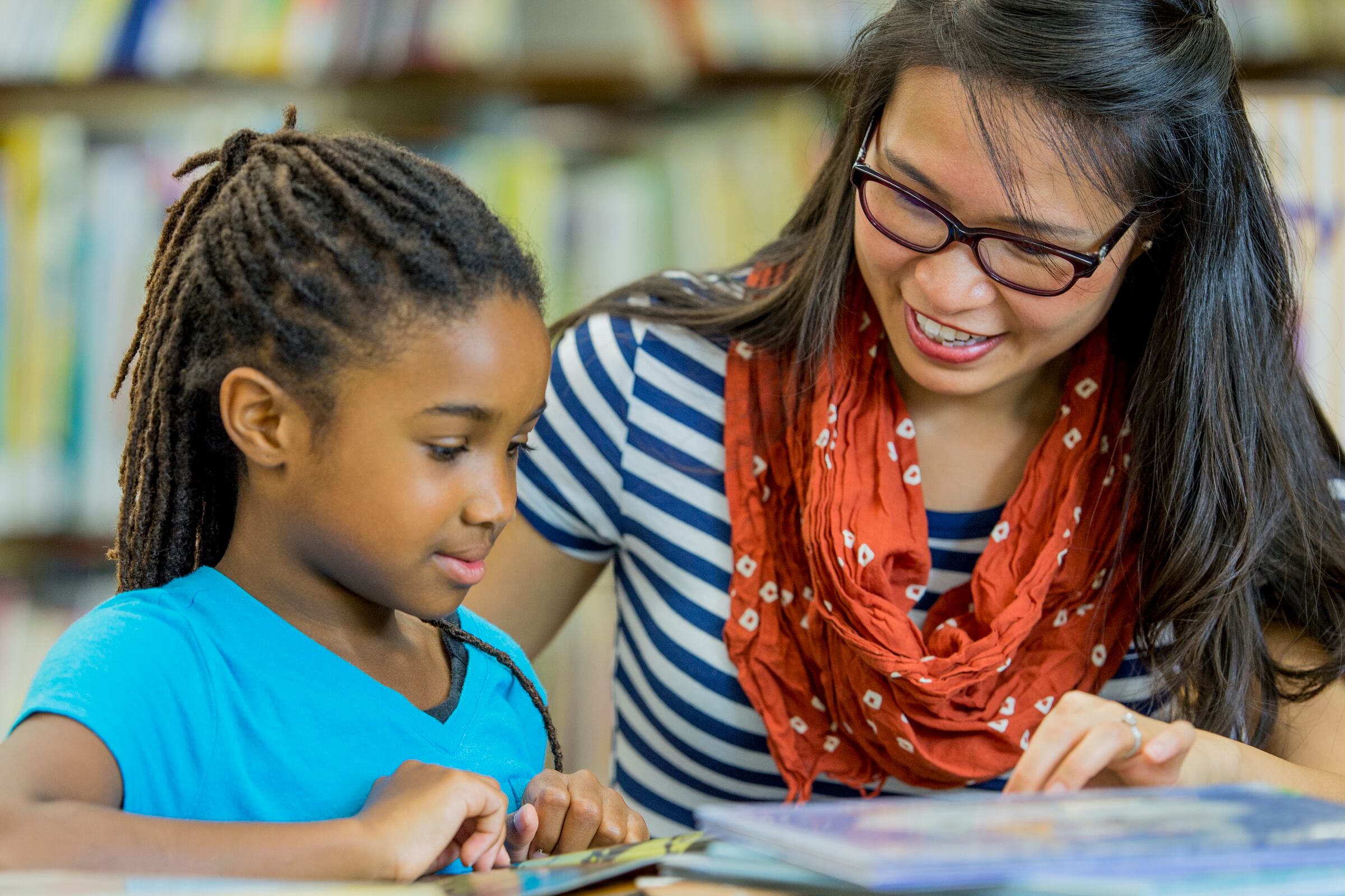 A teacher is reading books with an elementary age girl in the library.
