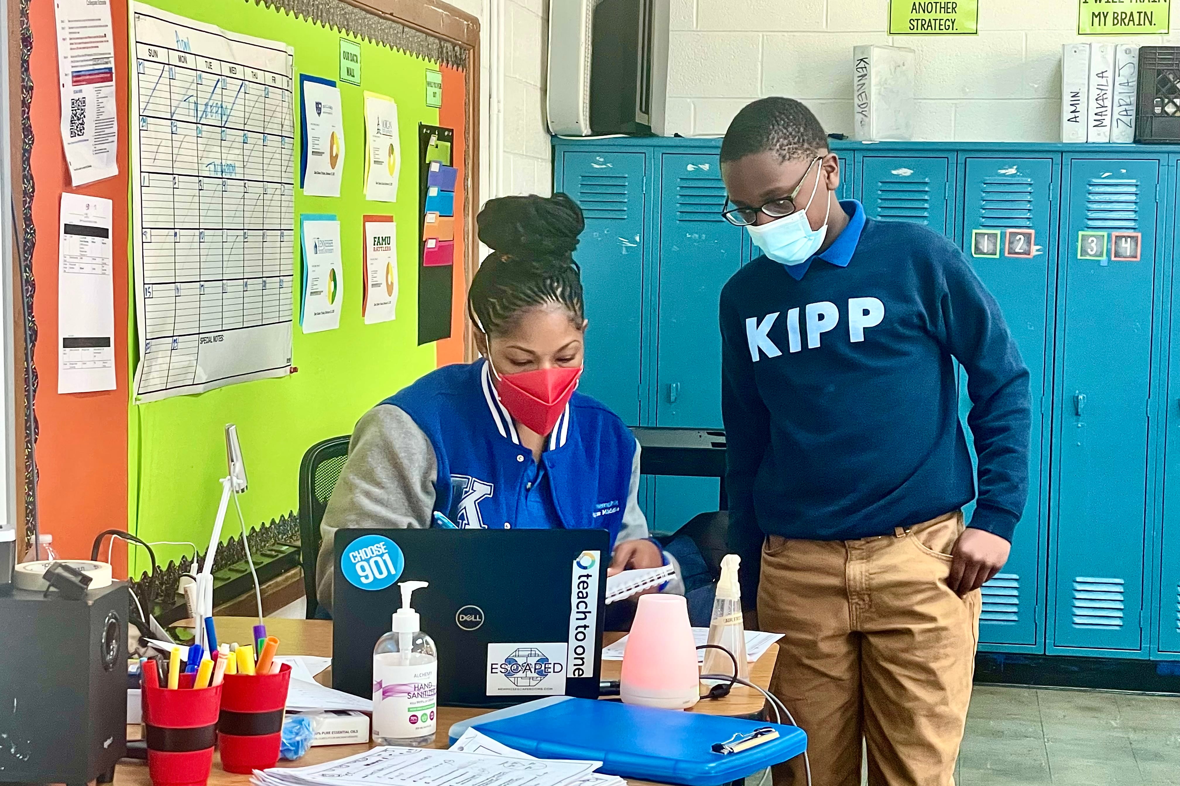 A female KIPP Memphis teacher answers a boy’s question while sitting at her desk in front of her laptop. Both are wearing masks; the teacher is wearing a bright blue and grey KIPP letter jacket, while the boy is wearing a navy sweatshirt with “KIPP” in brighter blue lettering.
