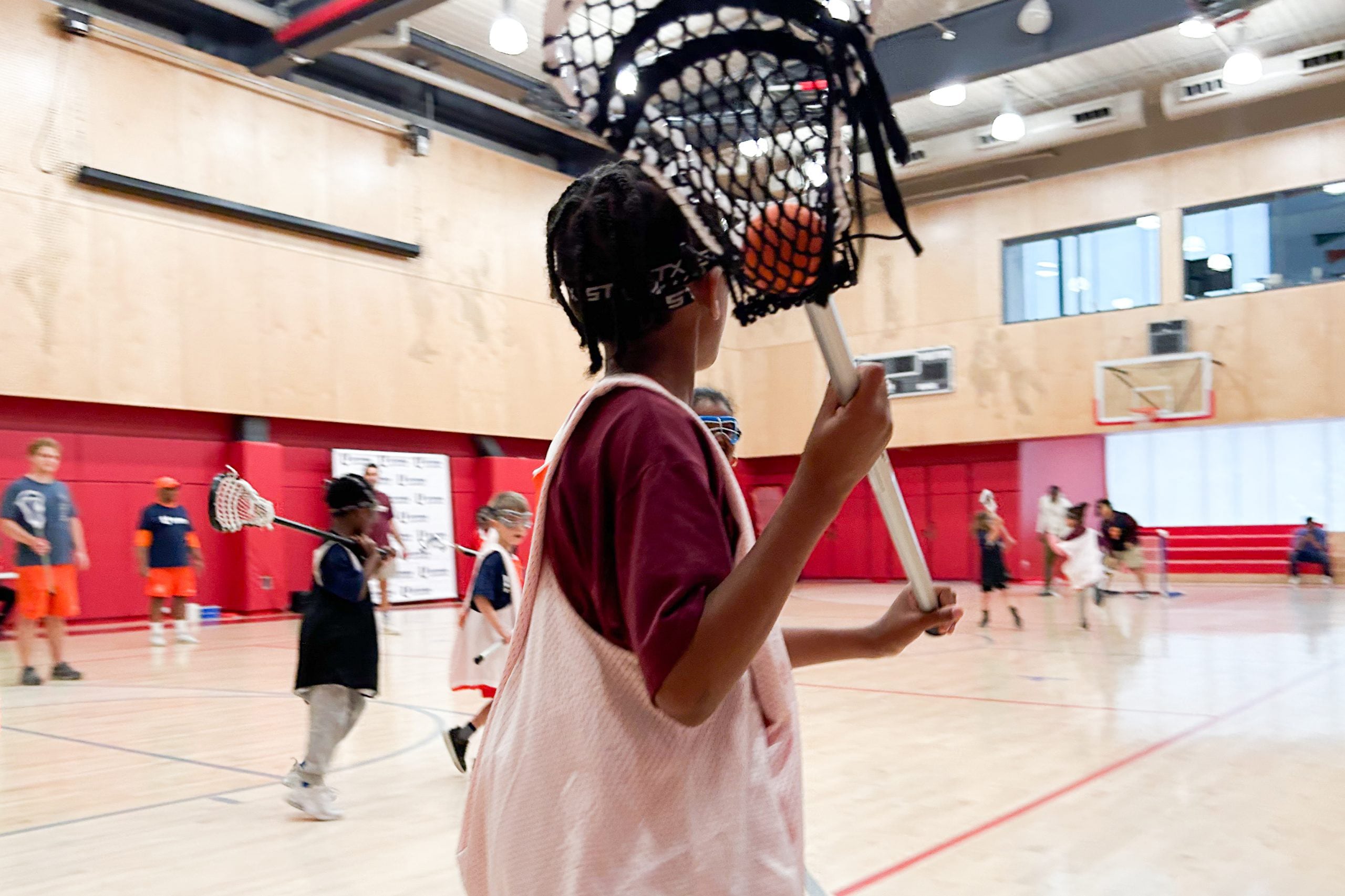 A young Black child holds a lacrosse stick with a ball in the net in a gymnasium with other children in the background.