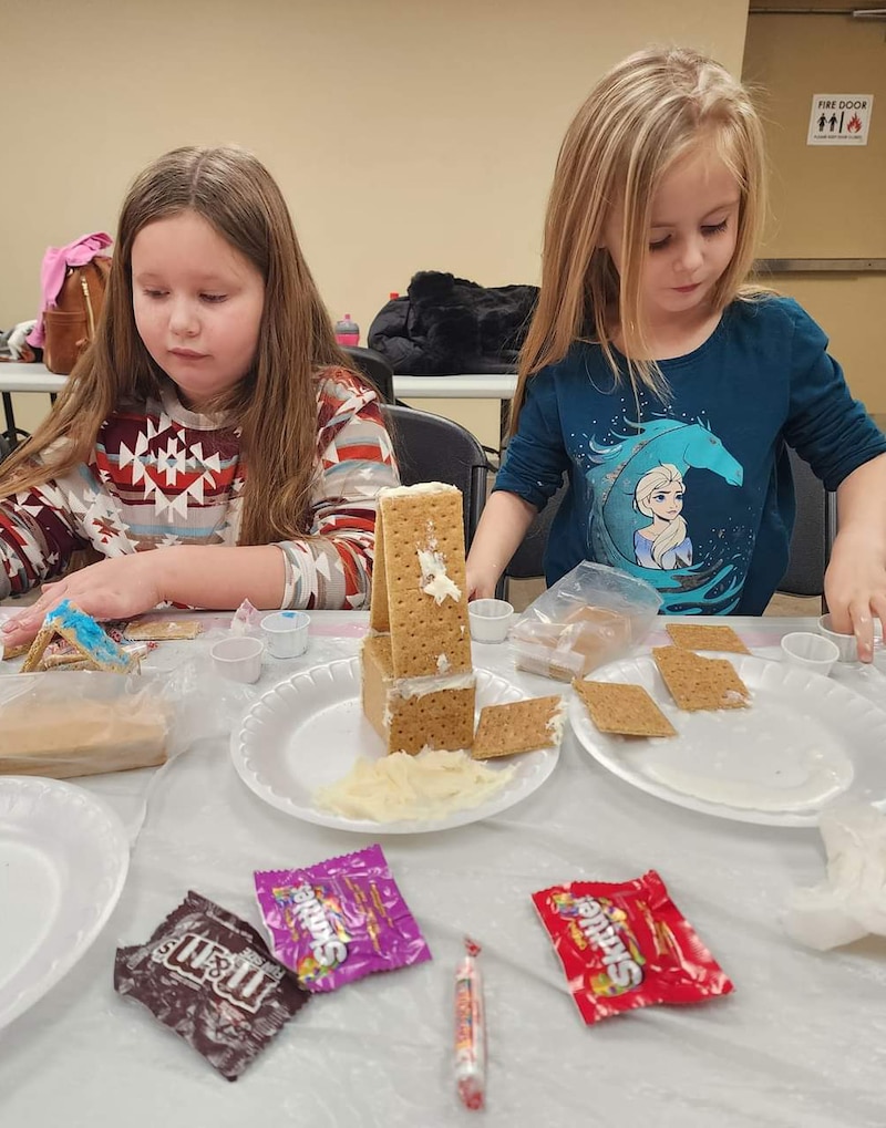 Two young children sit at a table making graham cracker ginger bread houses.