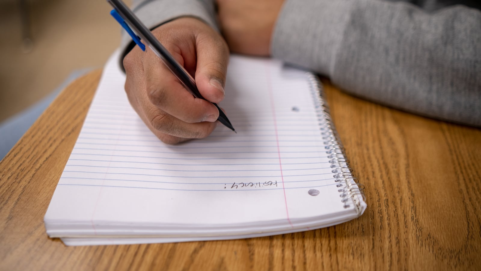 A young man writes in his notebook, using a mechanical pencil with a blue clip near the eraser.