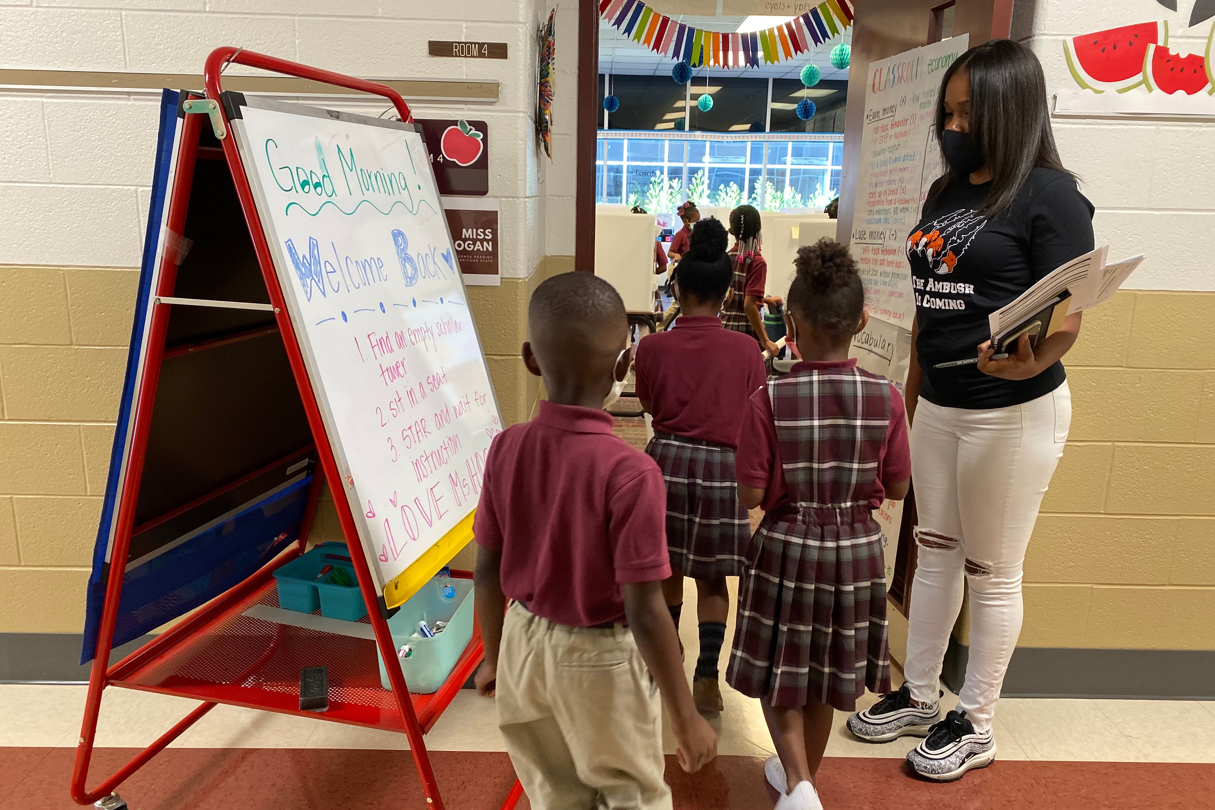 A line of elementary school students in uniforms walk past their teacher into their classroom.