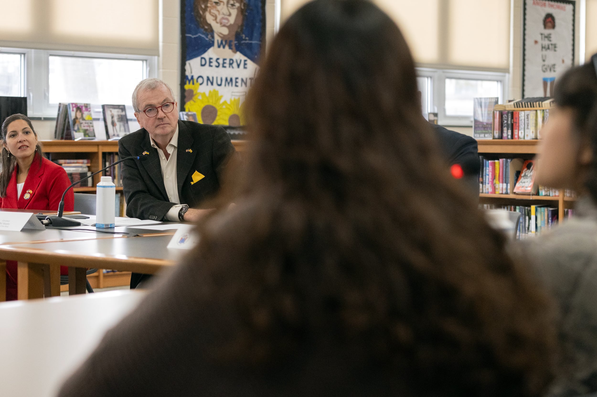 A man in a suit sits at a table in a school library with other people around him.