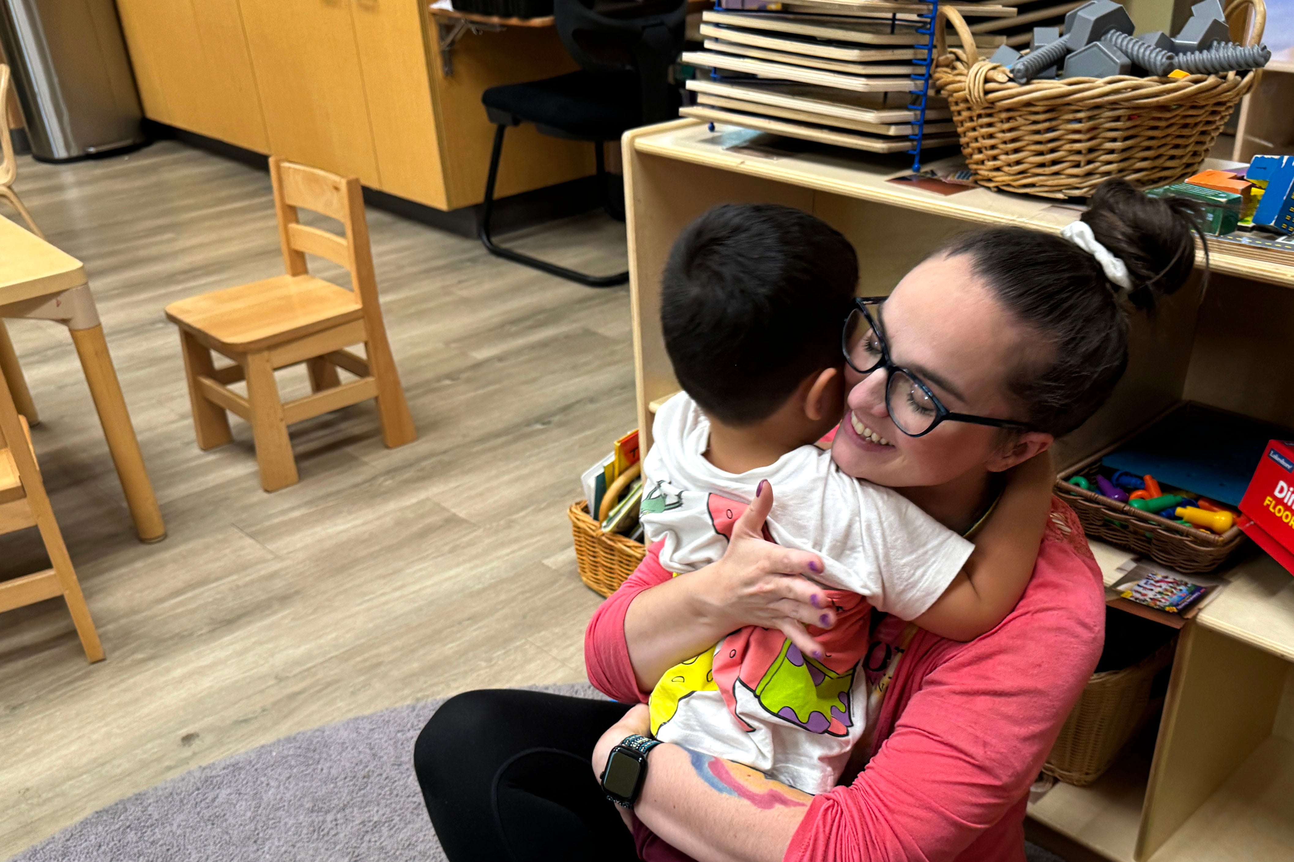 A young child hugs an adult woman wearing a pink shirt while sitting on the ground in a classroom.