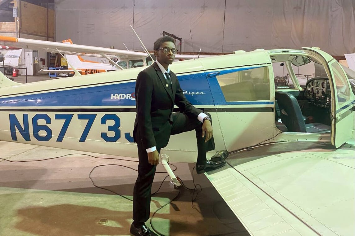 A high school student wearing a dark suit poses by the wing of a plane with a warehouse in the background.