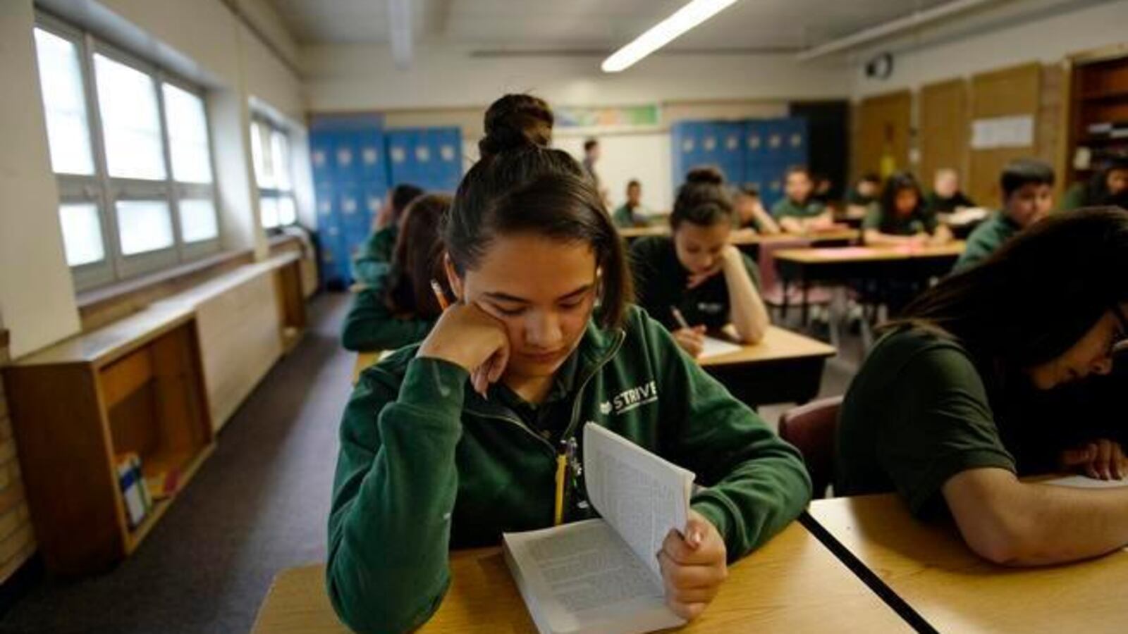 A girl with brown hair wearing a green shirt sits at a desk, reading a book.