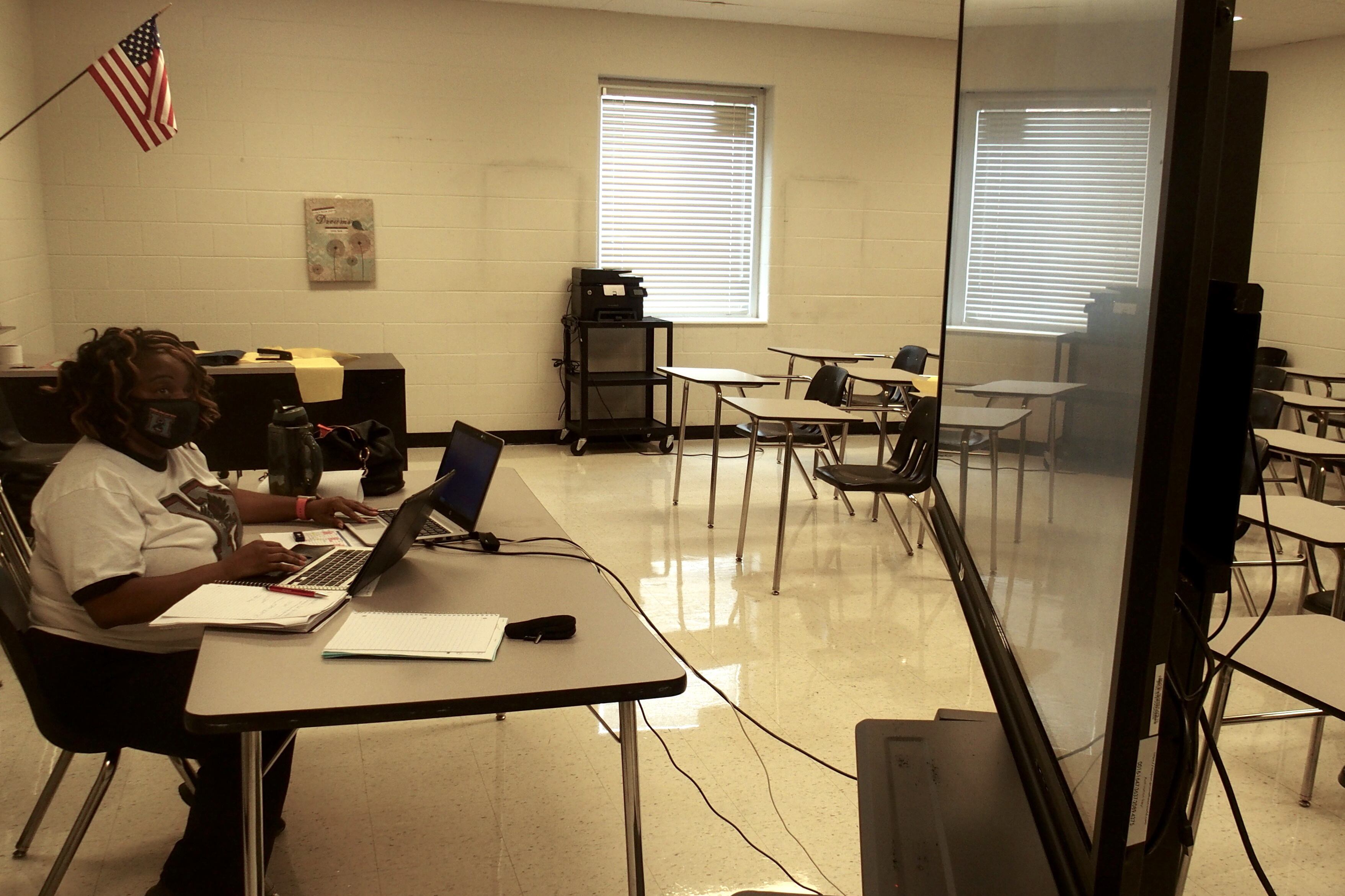 A teacher monitors the videoconference chat box on her laptop and a separate screen while another teacher conducts the class