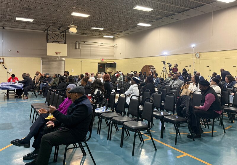 A photograph of a large group of people sitting in rows of chairs in a school gym.