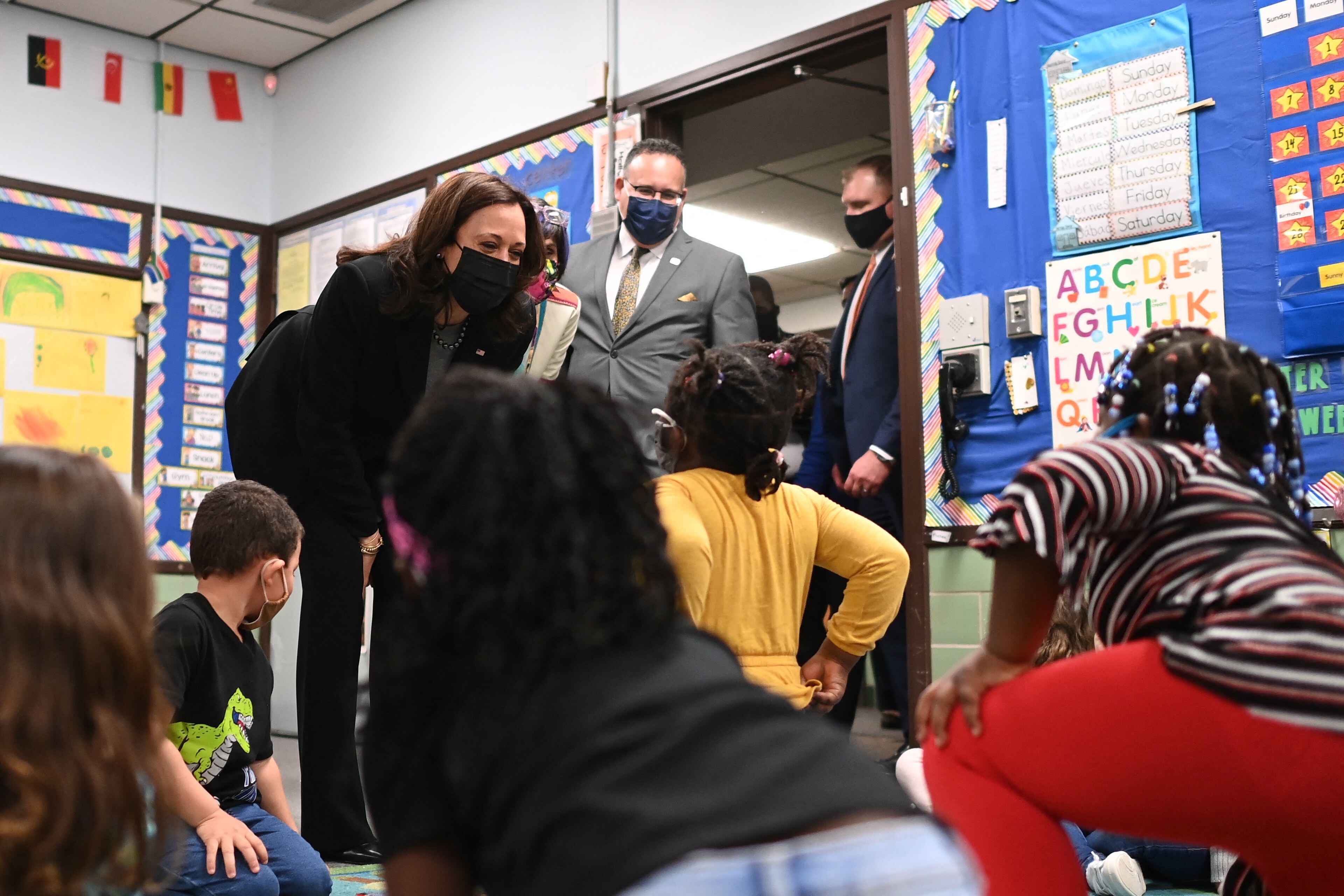 A classroom full of young children greet Vice President Kamala Harris, who is bending over to speak to the kids, as Secretary of Education Miguel Cardona stands behind her.