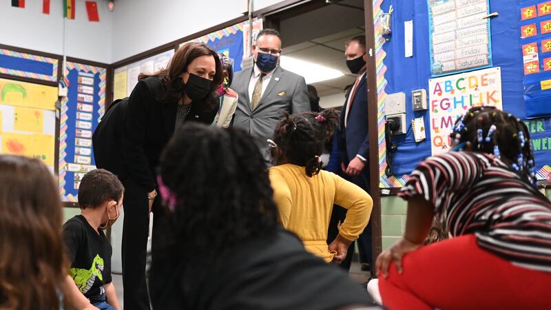 A classroom full of young children greet Vice President Kamala Harris, who is bending over to speak to the kids, as Secretary of Education Miguel Cardona stands behind her.