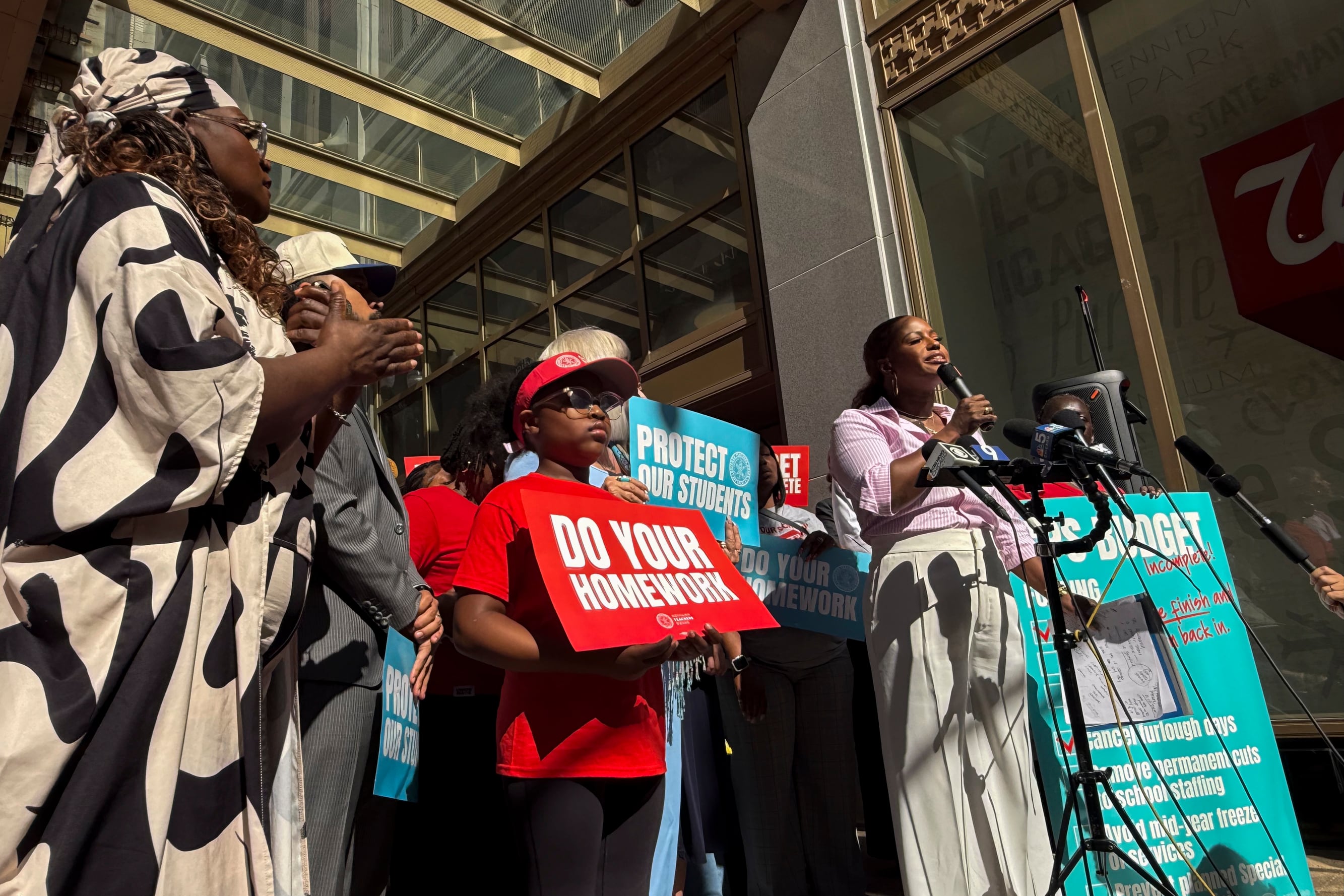 A Black woman is speaking into a microphone, with people surrounding her, holding signs that read "Do Your Homework."