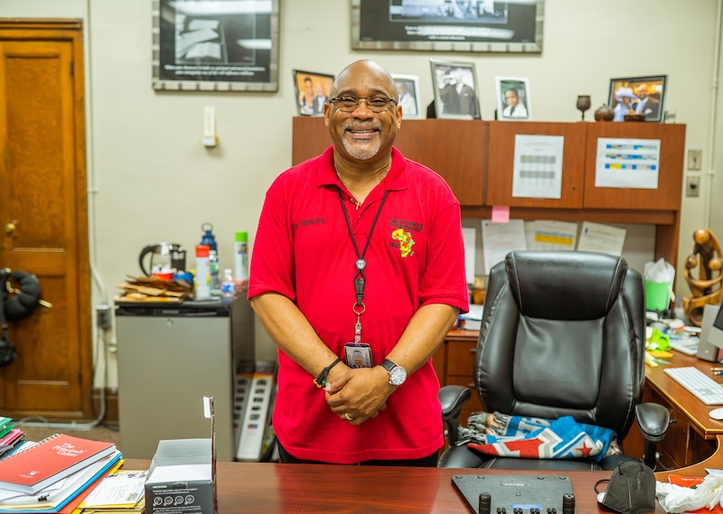 A man in a red shirt poses for a portrait standing at his desk in his office.