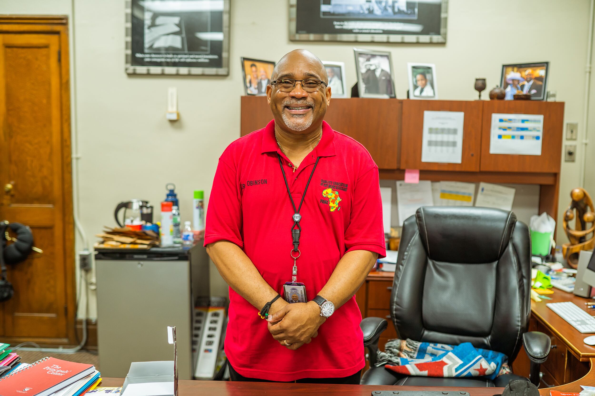 A man in a red shirt poses for a portrait standing at his desk in his office.