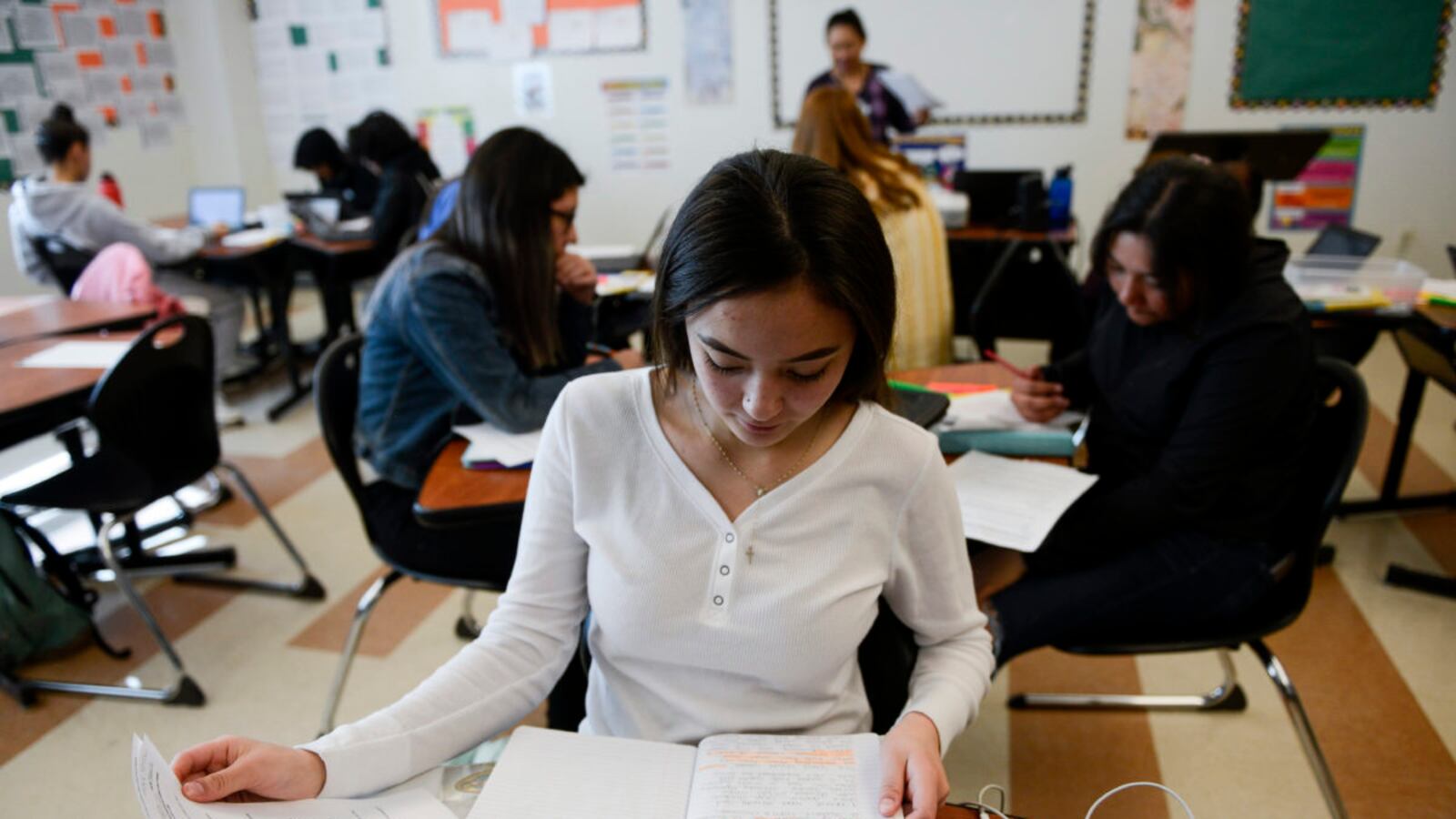 Adams City High School junior Alexandra Hernandez, in a white long-sleeve shirt, looks at her notebook, with a cell phone next to her. In the background are about five students working at tables and a teacher standing in front of a white board, in an AP language class on Feb. 4, 2019.